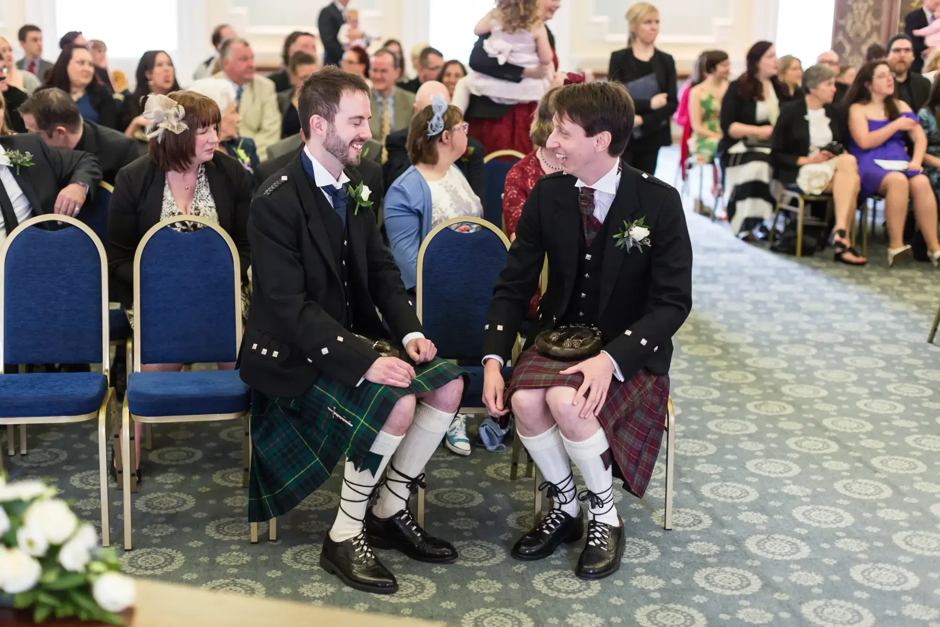 Two men in traditional scottish kilts and attire sitting and laughing together at a wedding ceremony, with guests seated around them.
