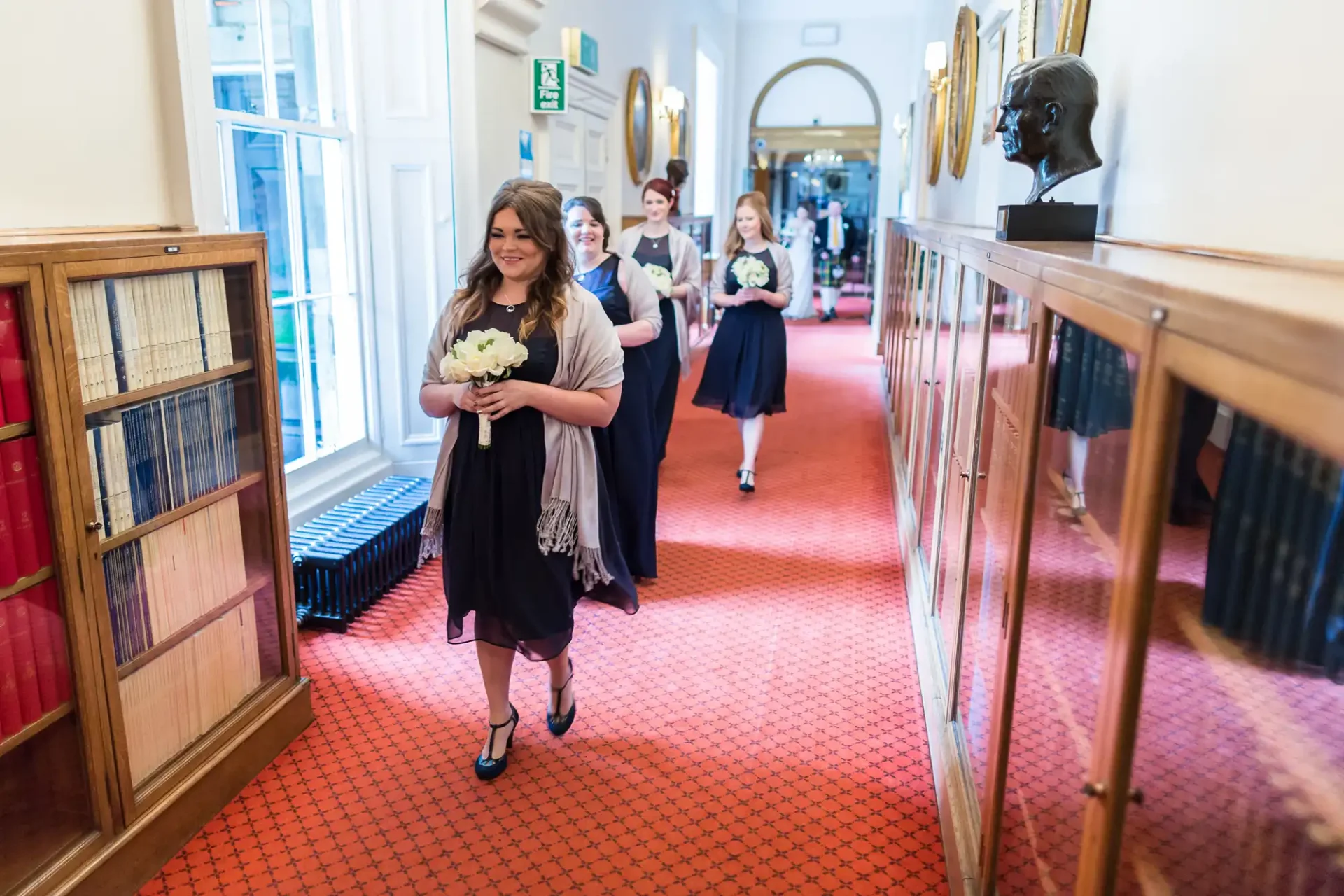 A woman leading a group of bridesmaids along a library hallway, all holding bouquets and dressed in formal attire.