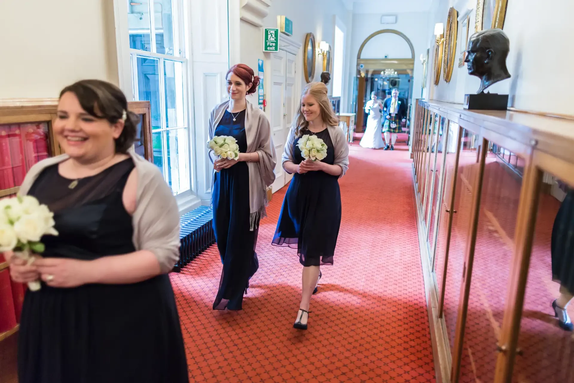 Three bridesmaids walking down a hallway, holding bouquets, and smiling during a wedding ceremony.