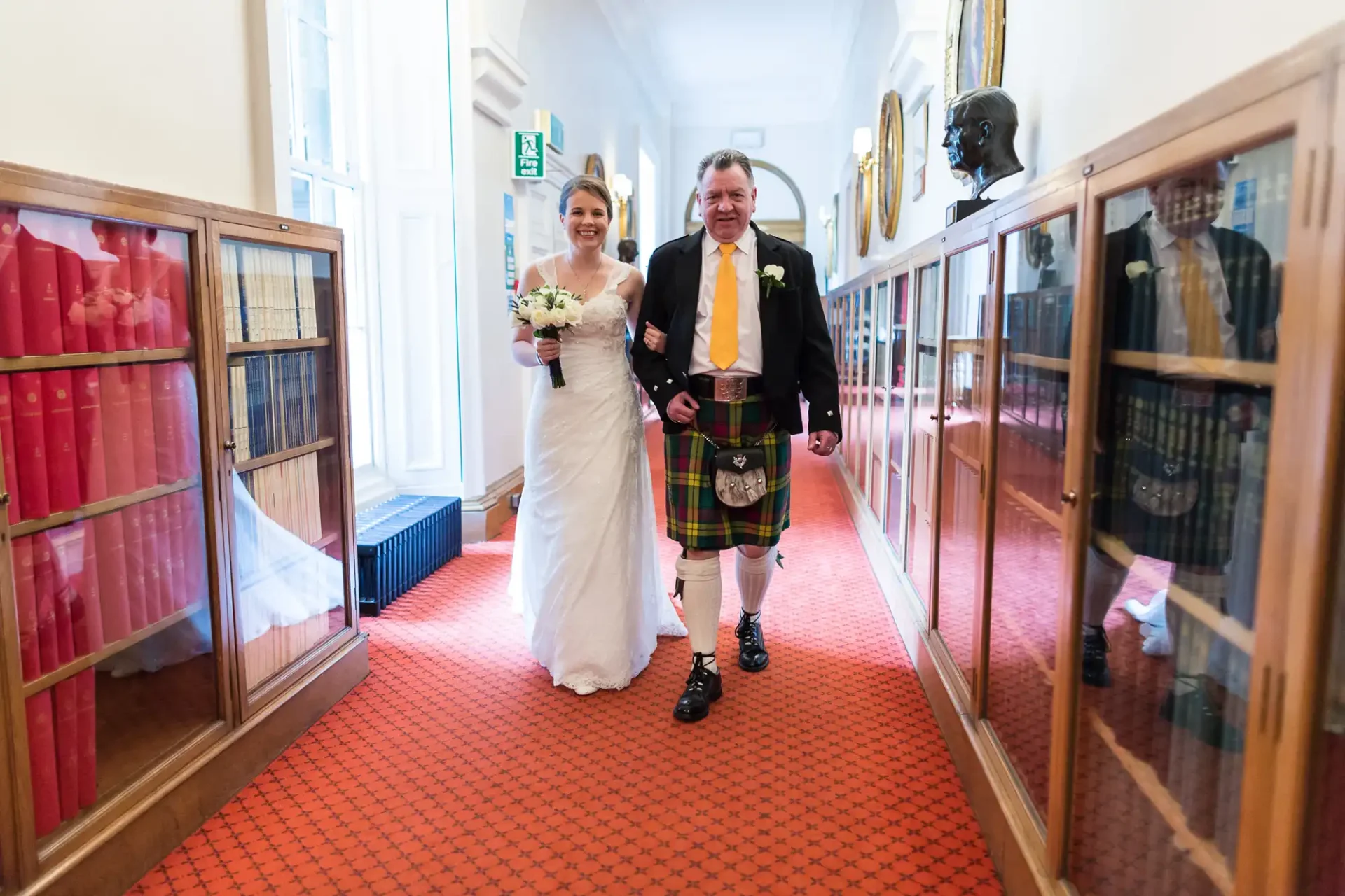 Bride in white dress and groom in scottish kilt walking down a hallway, smiling and holding hands, with display cases on either side.