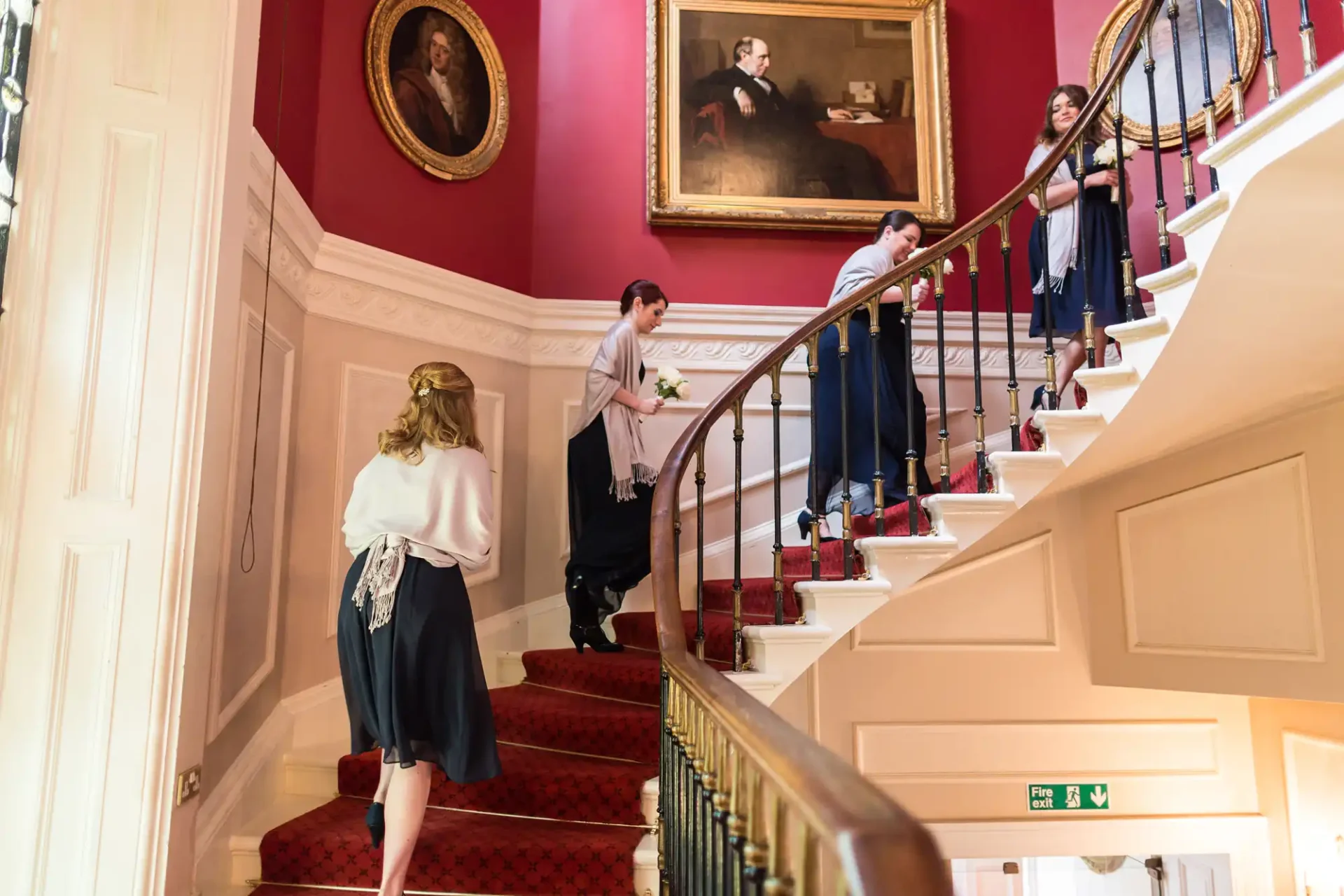 People ascending and descending a grand staircase with red carpet in an elegant room with pink walls and portraits.