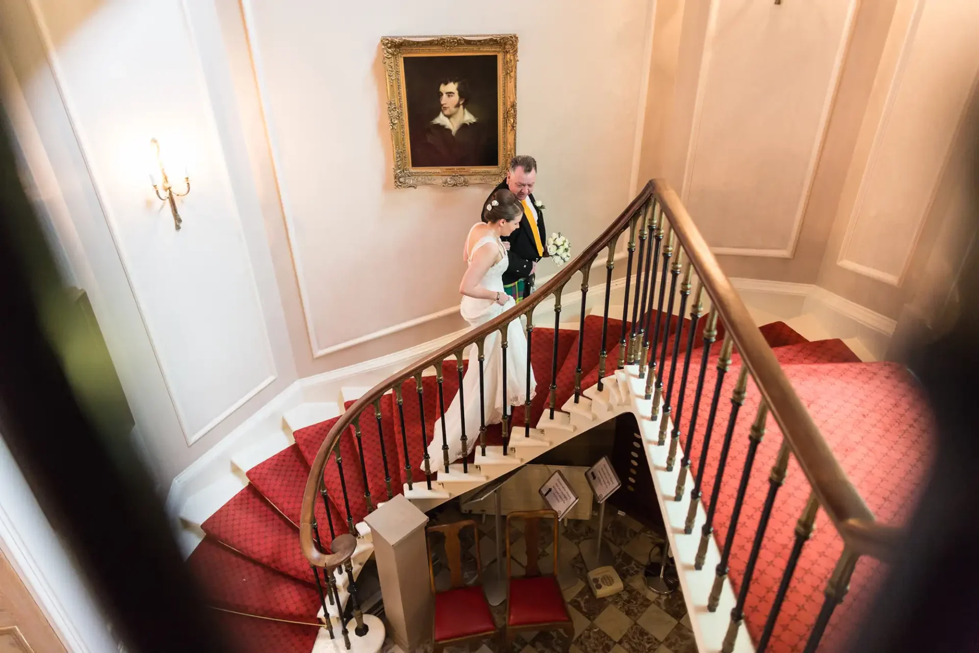 A bride and groom descend a curved staircase in an elegant hallway, with a portrait hanging on the wall above them.
