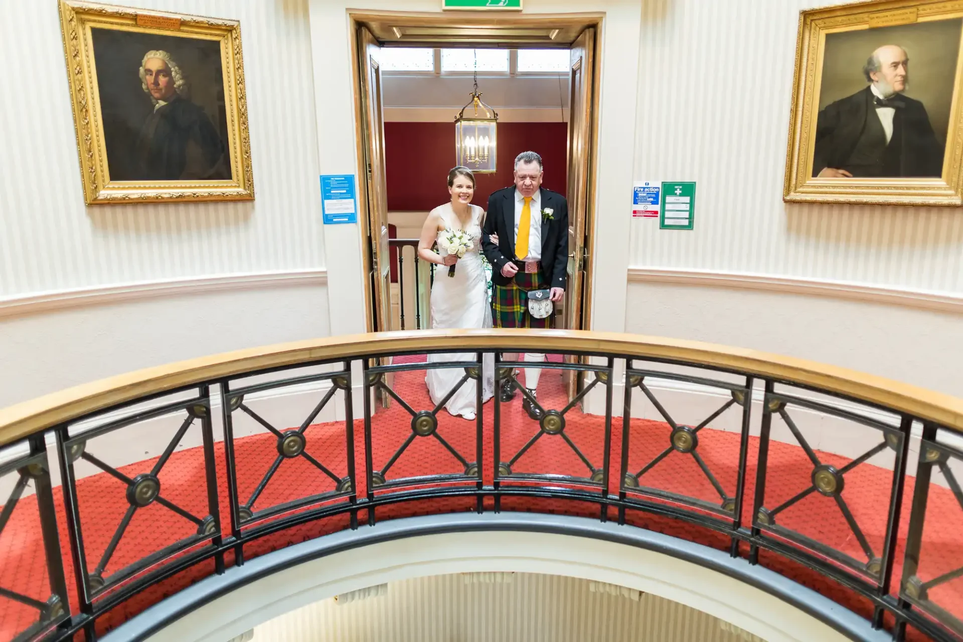 A bride and groom holding hands on a curved staircase in a hotel, with portraits hanging on the walls.