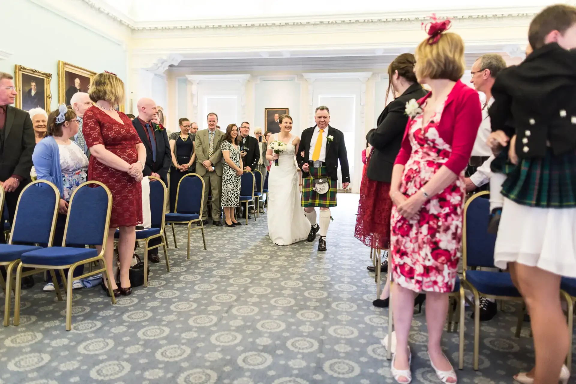 Bride and groom walk down the aisle, surrounded by guests, in a room with blue chairs and ornate carpet. the groom wears a kilt.