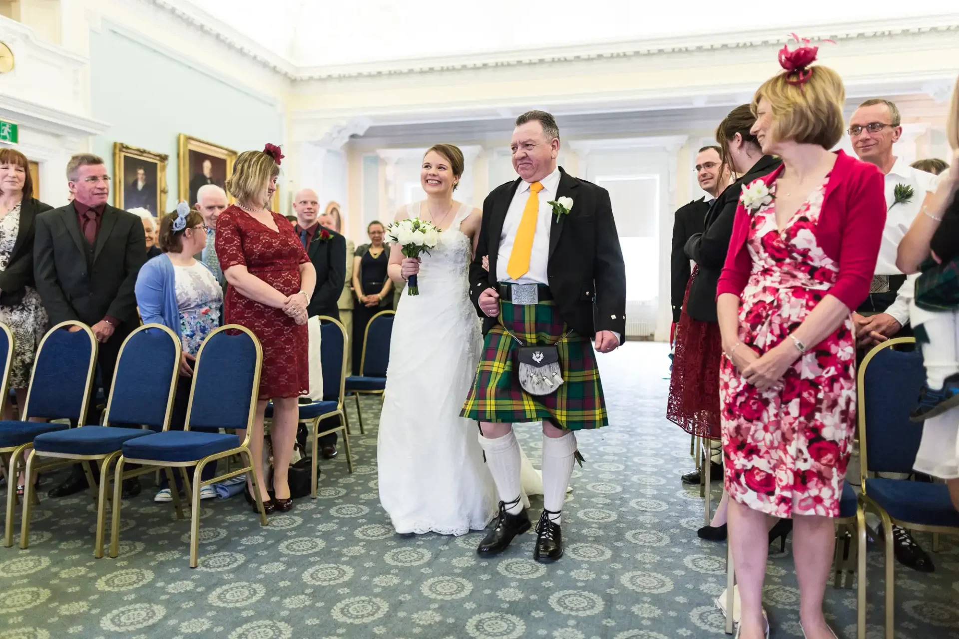 Bride and groom walking down the aisle, the groom in a traditional kilt, surrounded by smiling guests in a decorated room.