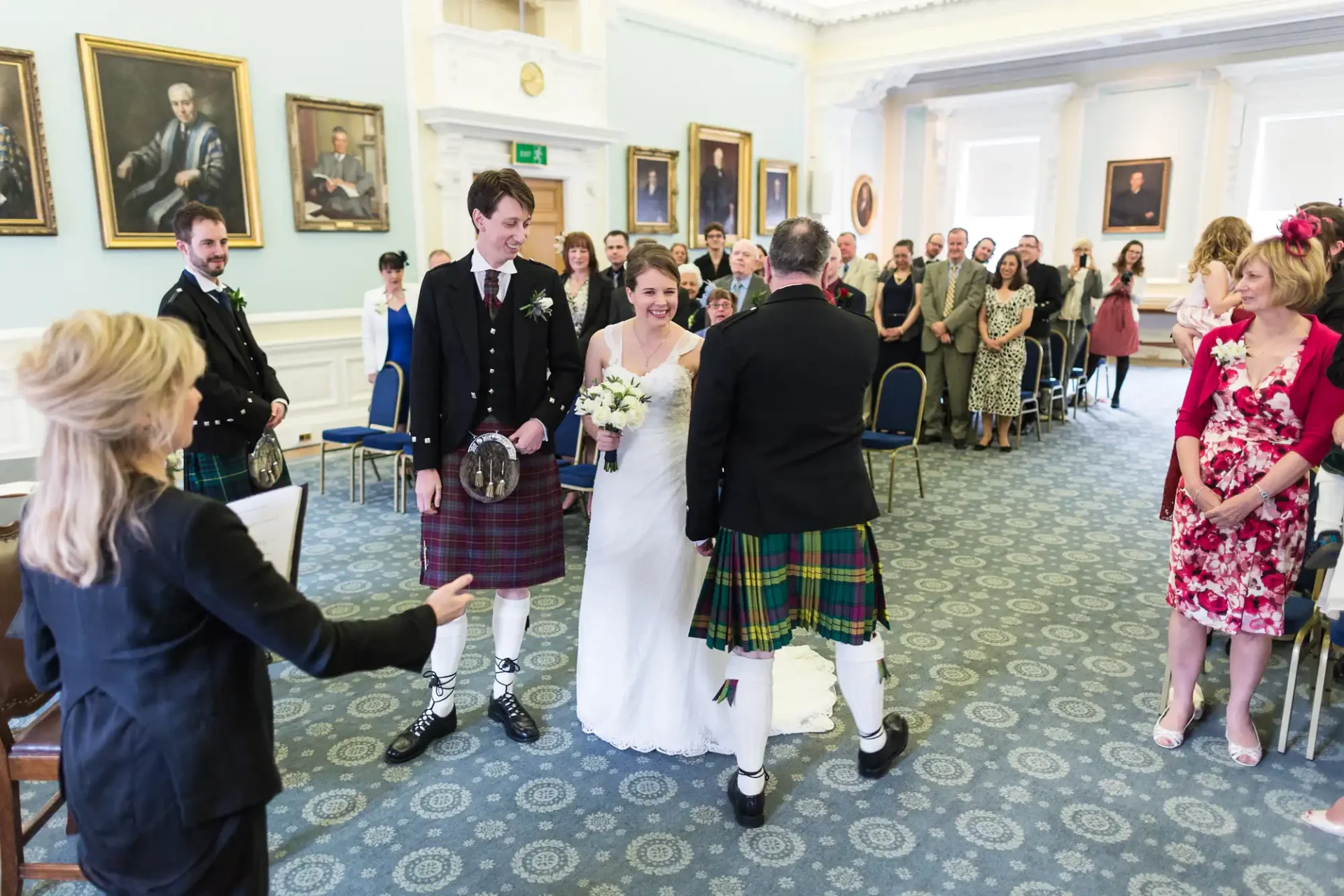 A joyful bride and groom walk down the aisle, the groom in a kilt, as guests watch and a woman scatters flower petals in a decorated room with portraits on the walls.