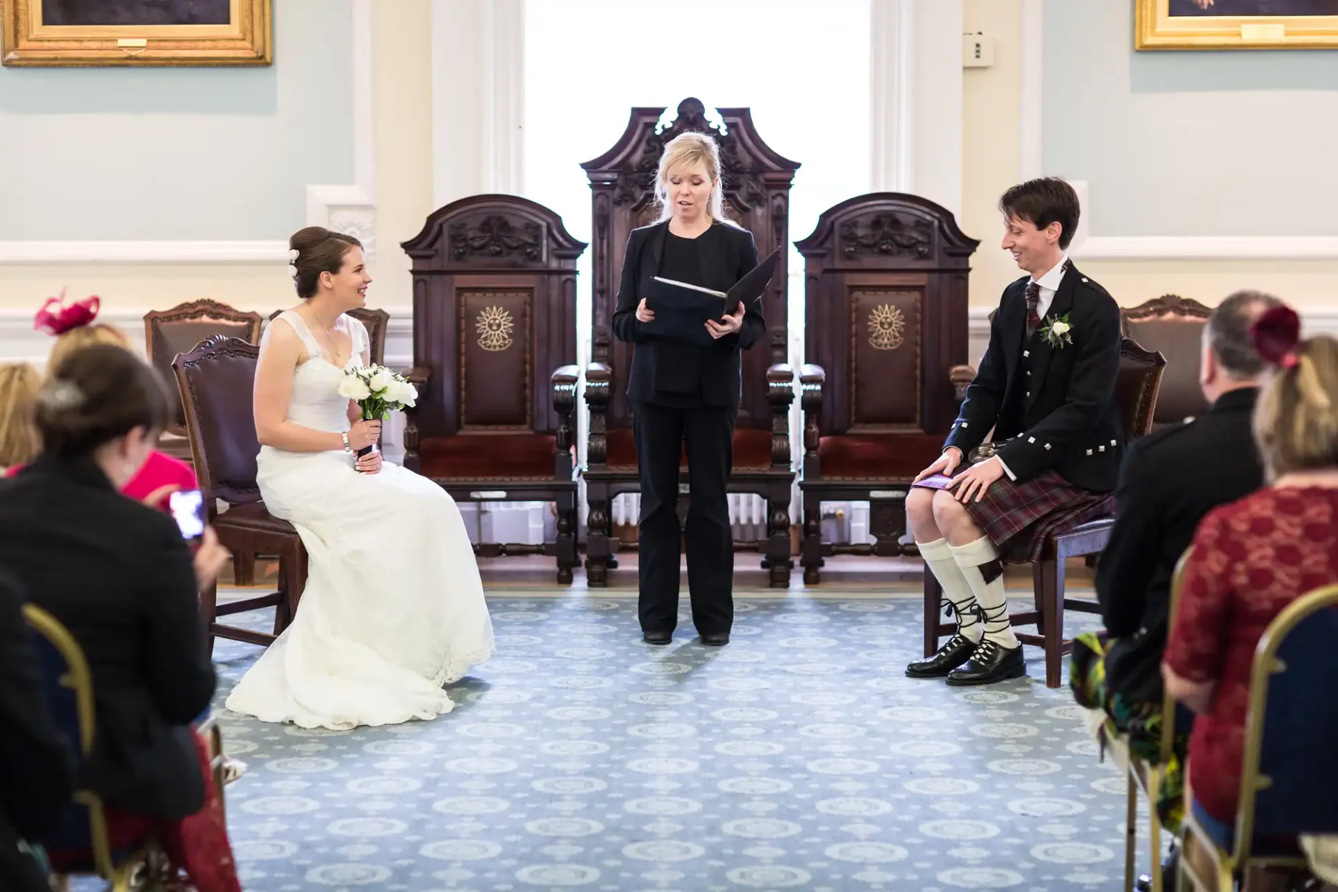 Bride in white dress and groom in scottish kilt sit facing each other at a wedding ceremony, with an officiant speaking and guests seated around them.