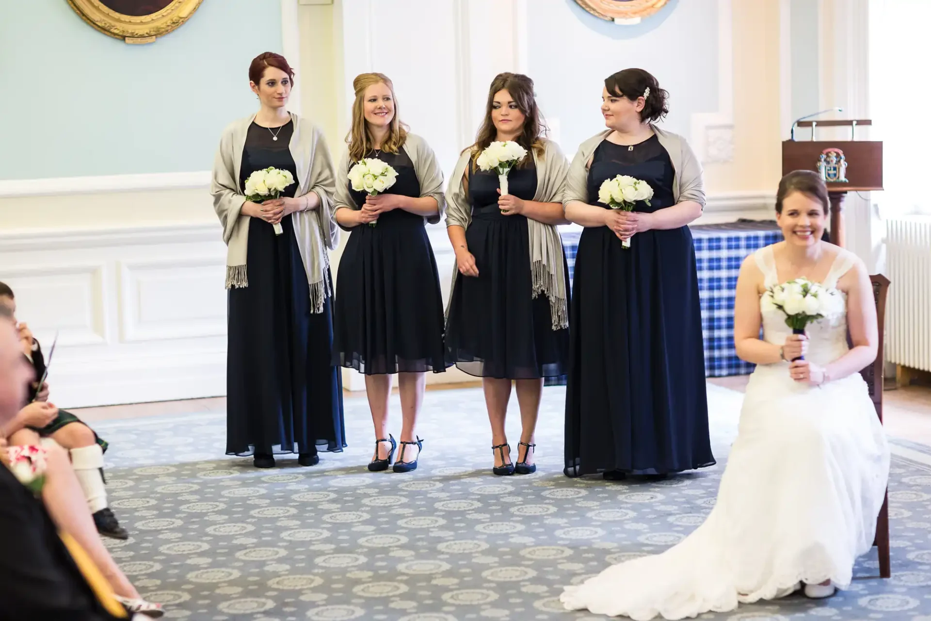 Four bridesmaids in navy dresses and a bride in white at a wedding ceremony, all holding bouquets, smiling as they watch something off-camera.