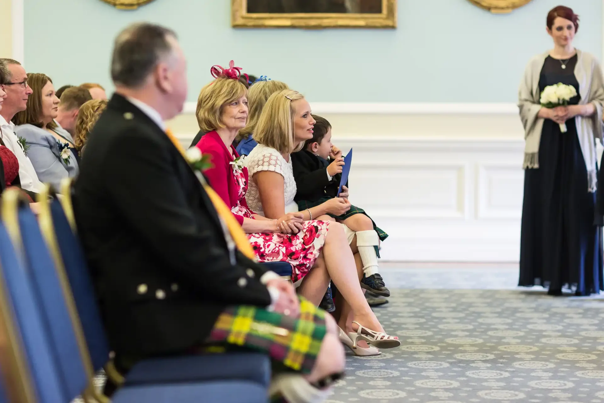 A group of guests seated in rows at a formal event, attentively watching a ceremony. a man in a kilt and a woman holding a bouquet are visible.