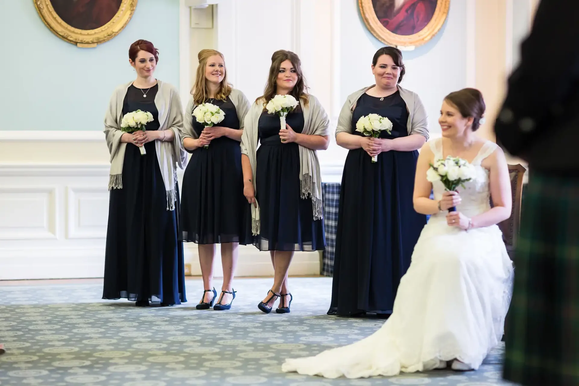 Four bridesmaids holding bouquets stand in a line, smiling towards a bride seated in the foreground in a wedding hall.