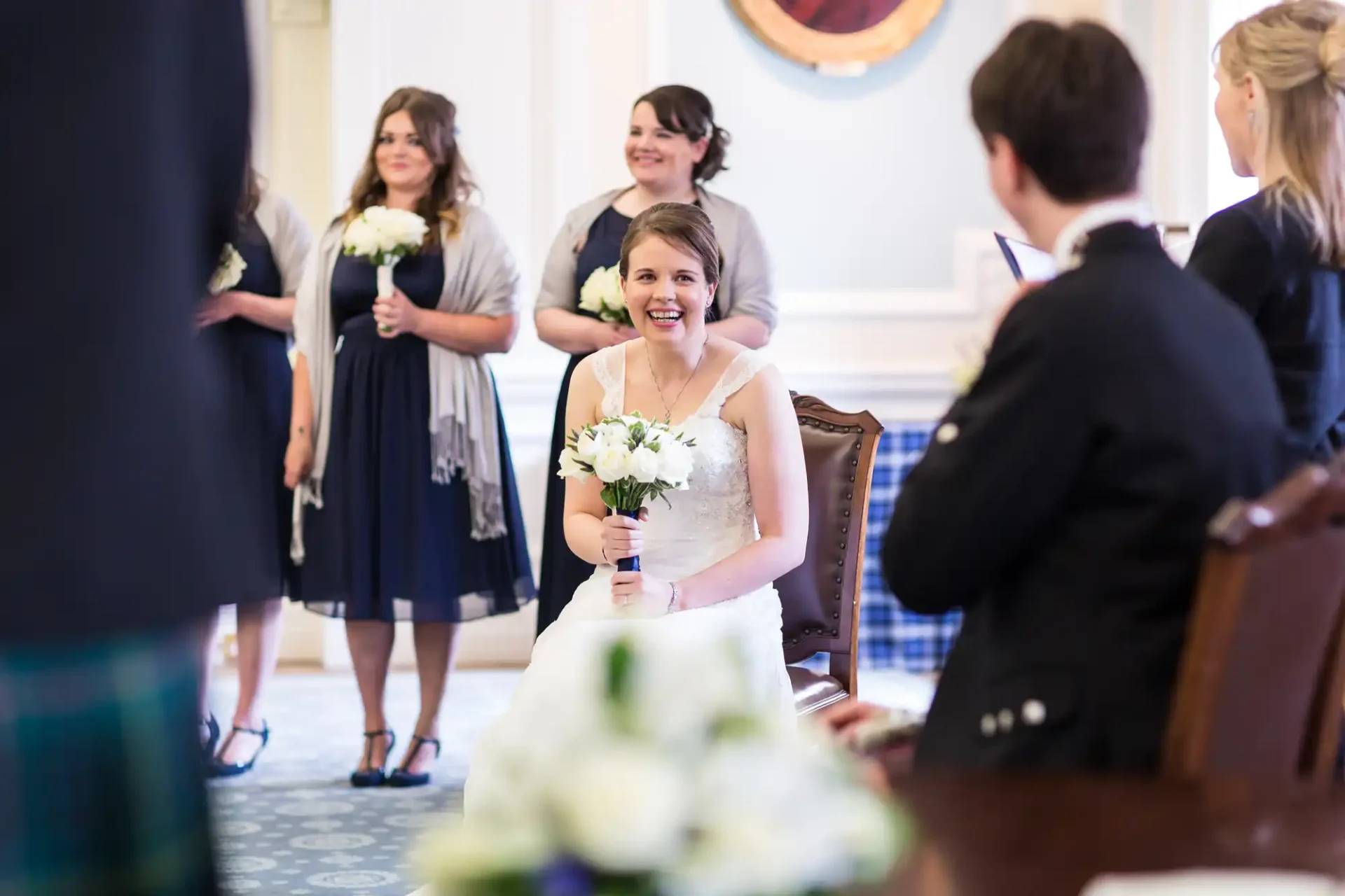 A bride in a white dress holding a bouquet smiles while seated, surrounded by bridesmaids and guests in a ceremonial room.