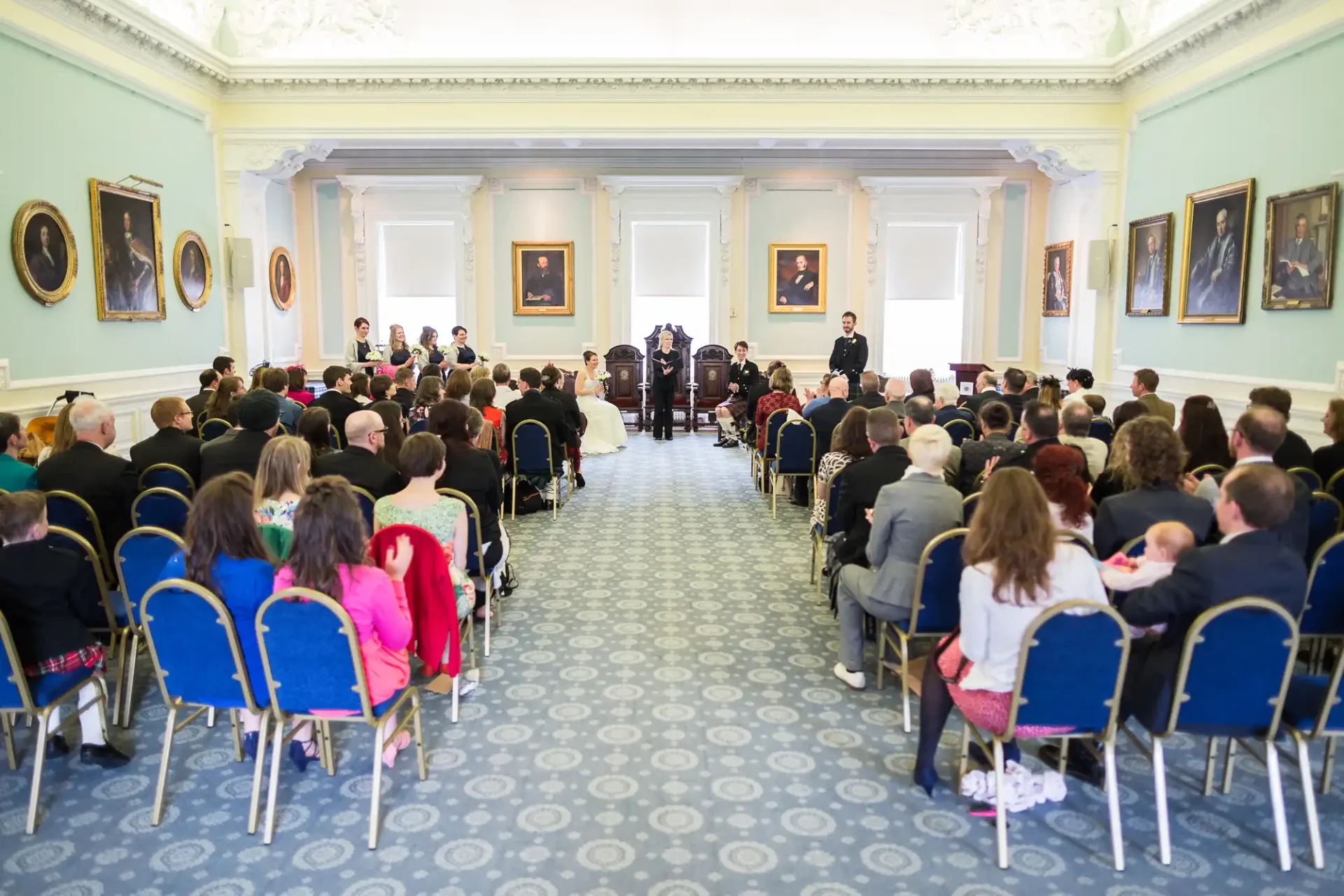 Wedding ceremony in an elegant room with seated guests, bride and groom at the front, and portraits on the walls.