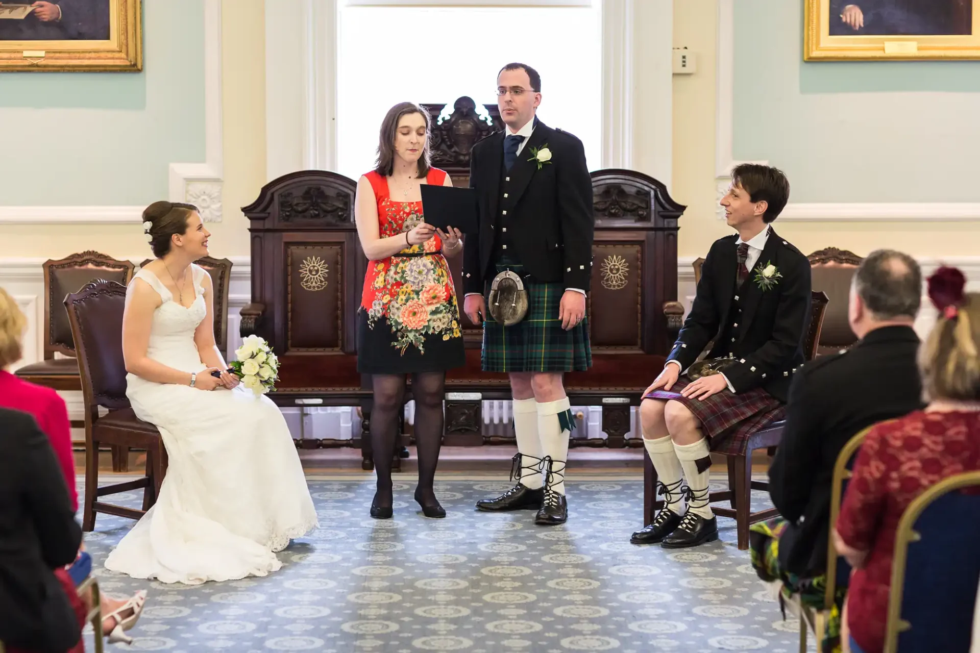 A wedding ceremony in a room with a woman in a floral dress and a man in a kilt standing at the front, while another woman sits and listens.