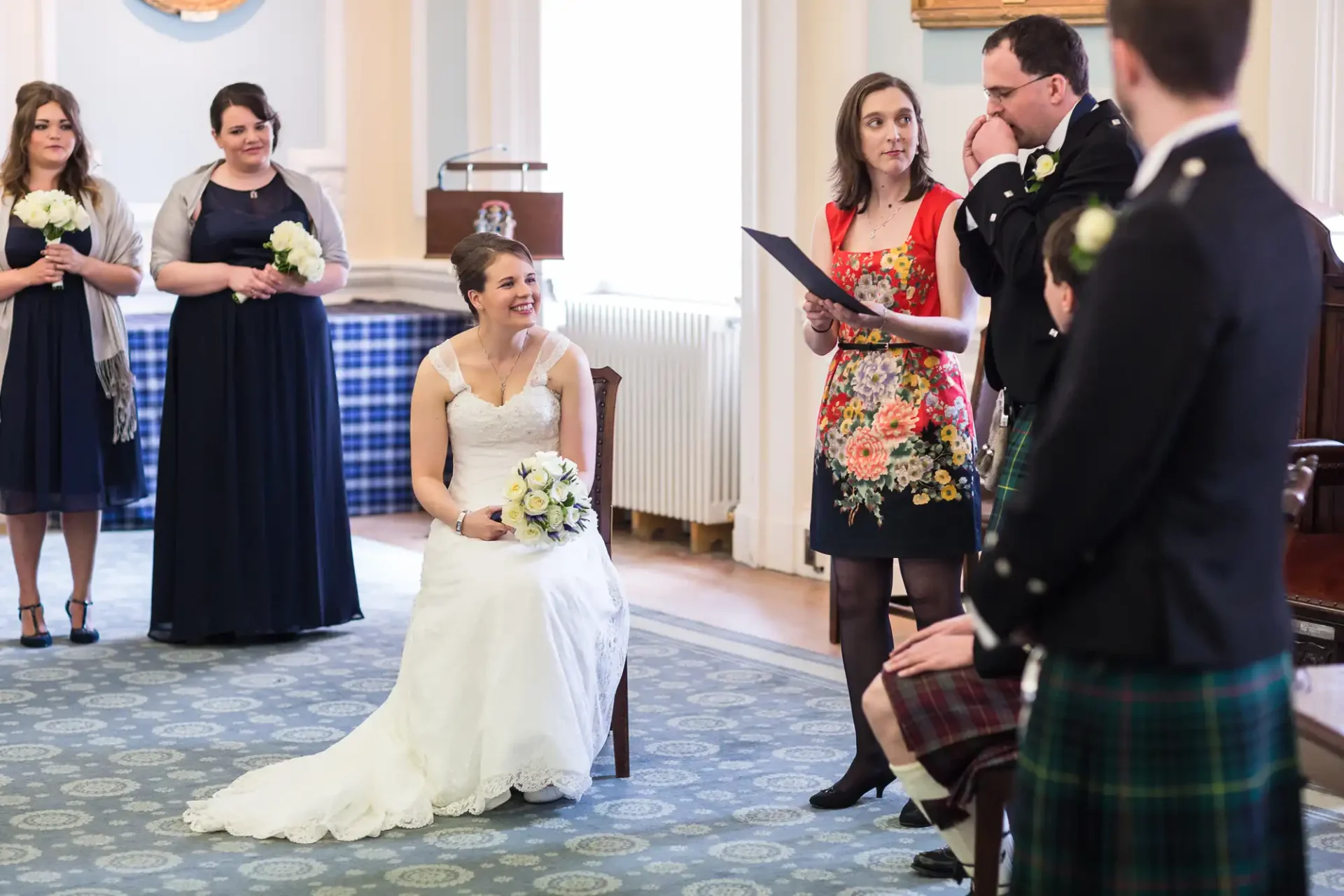 A bride seated, laughing joyfully at her wedding ceremony, with groom, officiant, and bridesmaids nearby in a room with elegant decor.