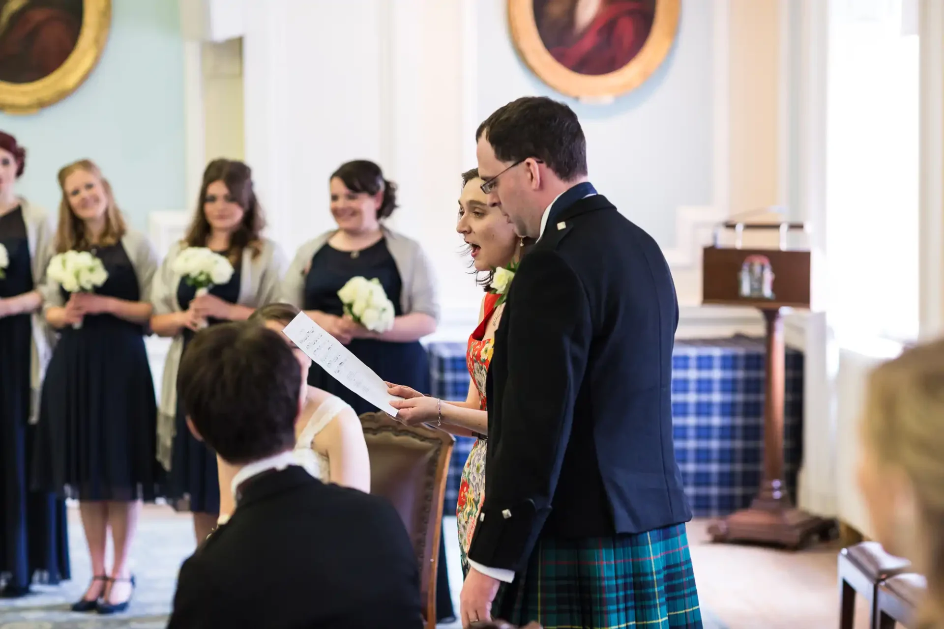 A couple reads from a paper during their wedding ceremony, standing before a small group of guests in a room with portraits on the walls.