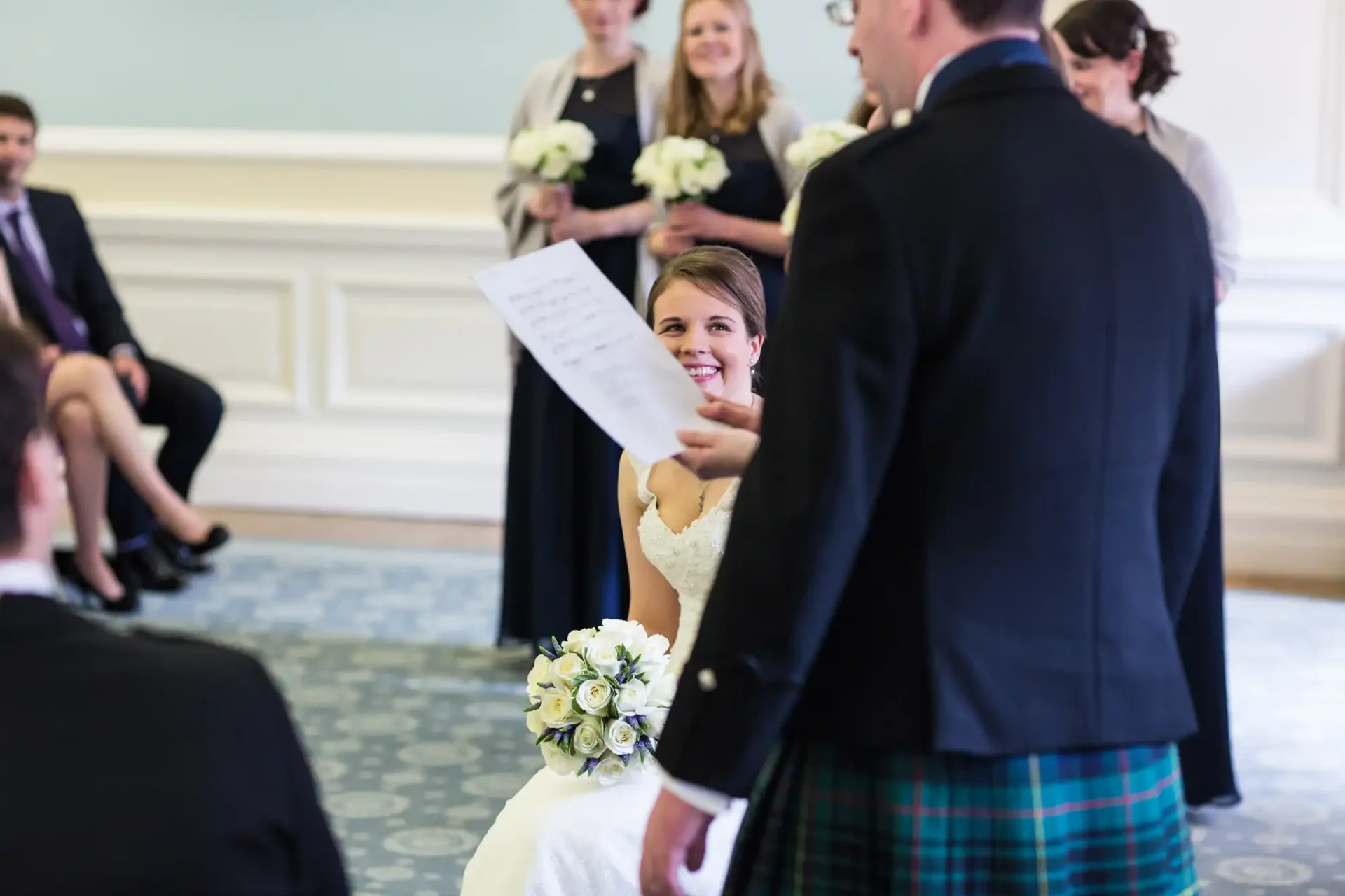 A bride in a white dress smiling and looking up at a man in a kilt during a wedding ceremony, with guests and bridesmaids in the background.