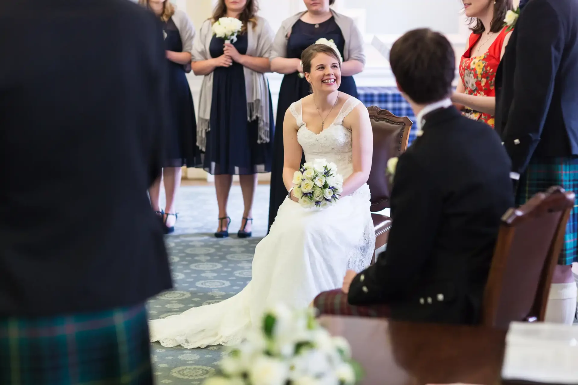 A bride in a white dress laughs joyfully while seated, facing a groom in a kilt, surrounded by guests in a room with floral decorations.