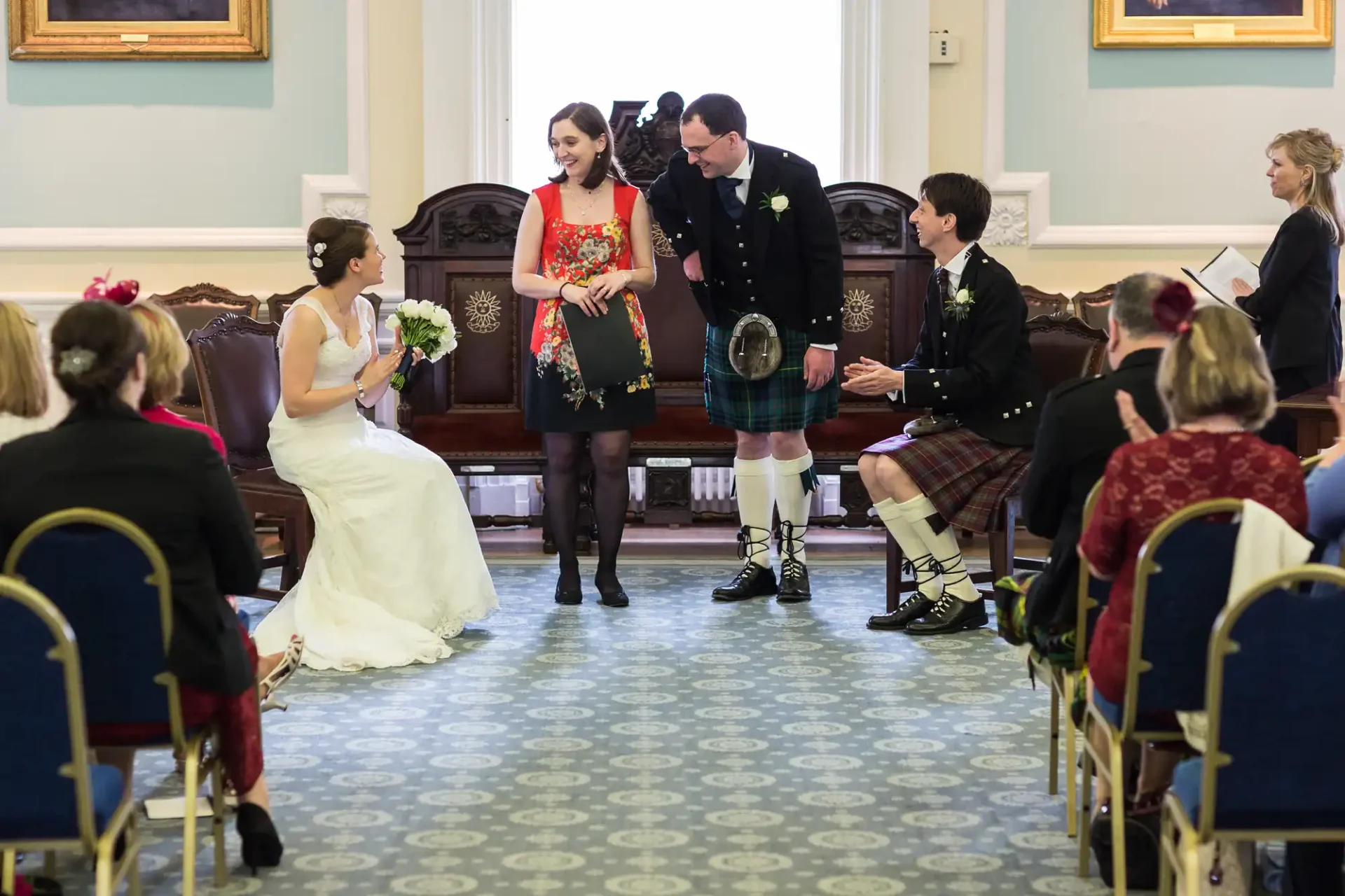 A bride in a white dress and a groom in a kilt smile at each other, holding hands in a hall with seated guests.