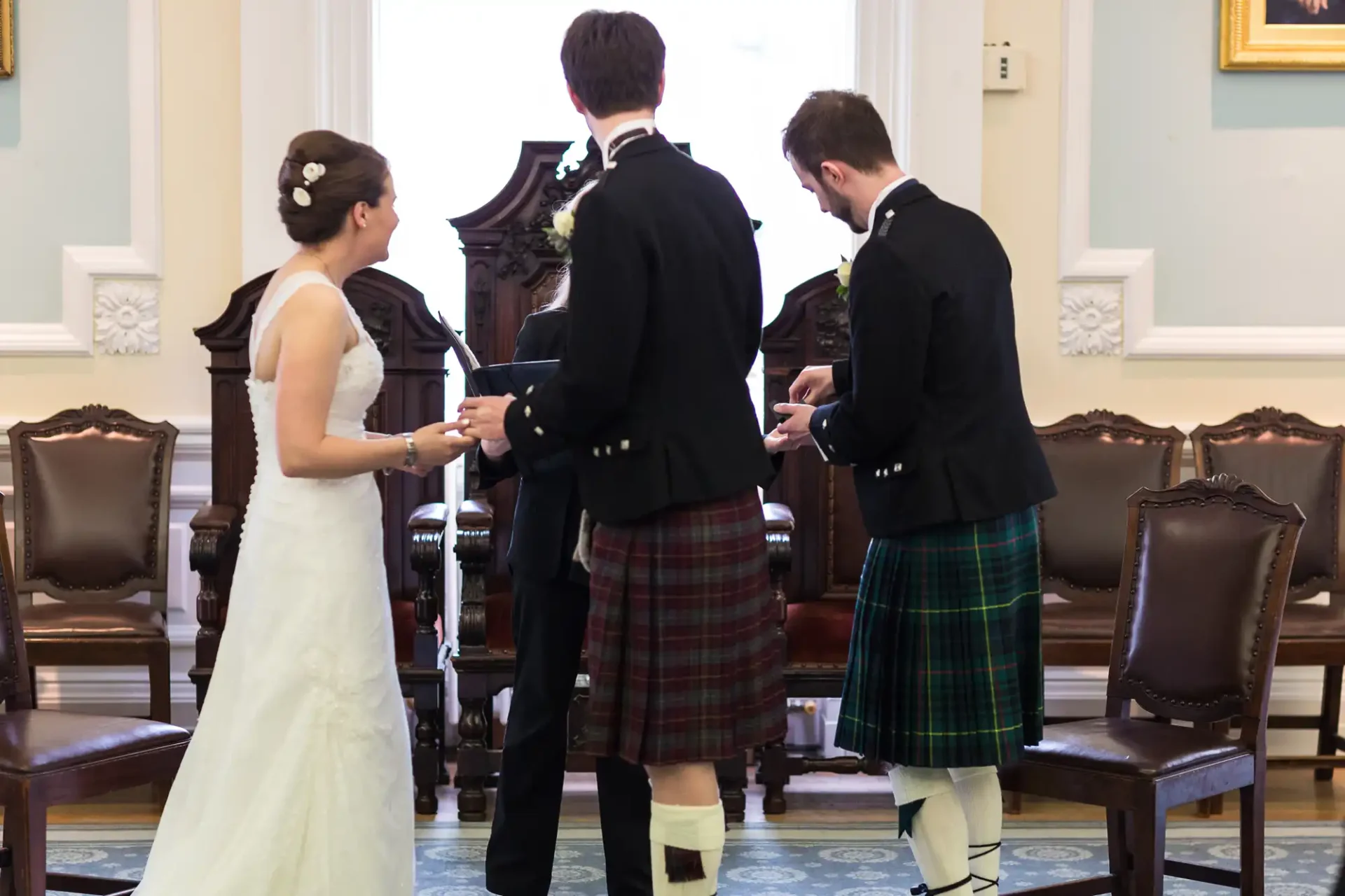 A bride and two men in kilts during a wedding ceremony inside an elegant room, facing away from the camera.
