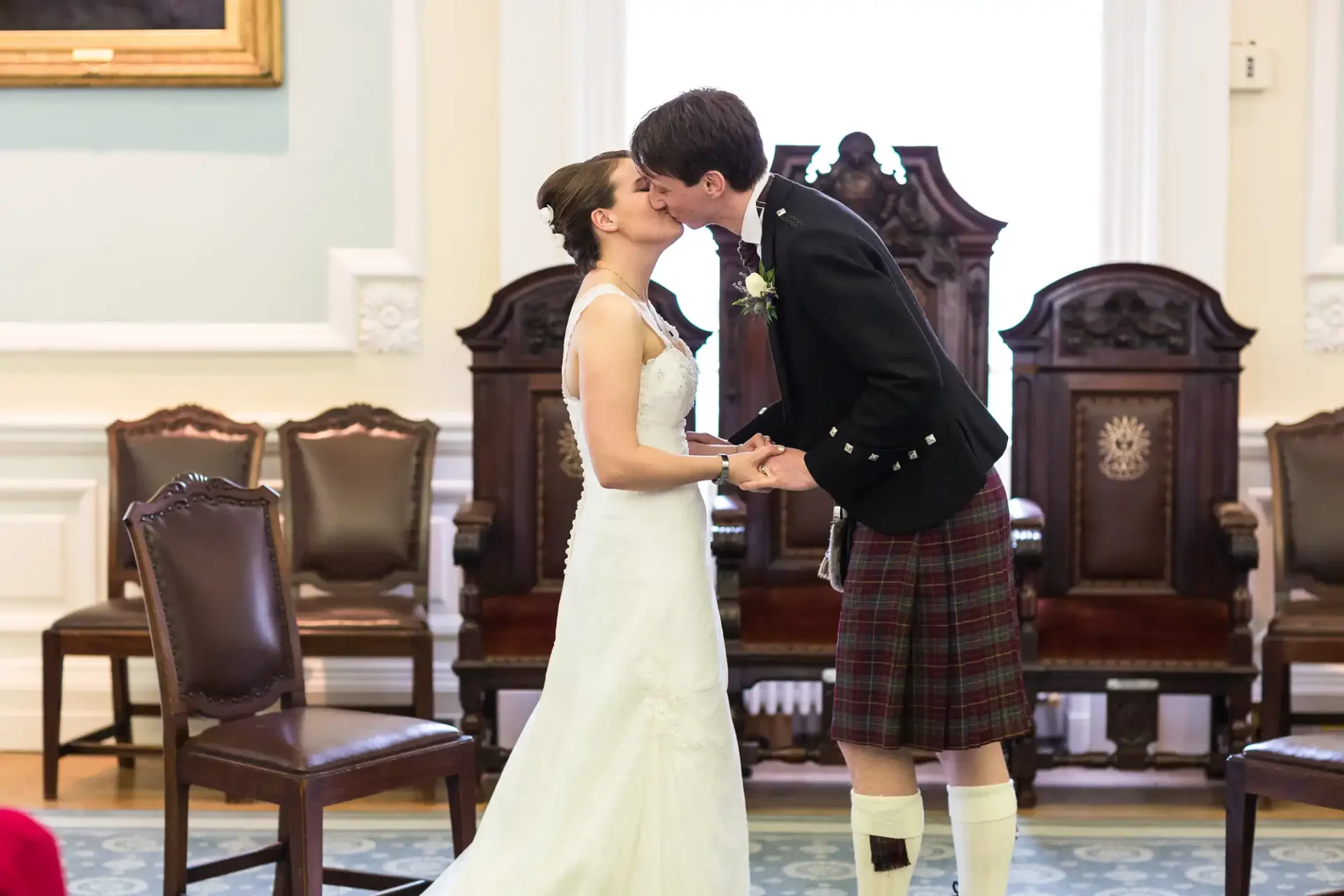 A bride and groom kiss during their wedding ceremony in an elegant room, the groom wearing a traditional kilt.