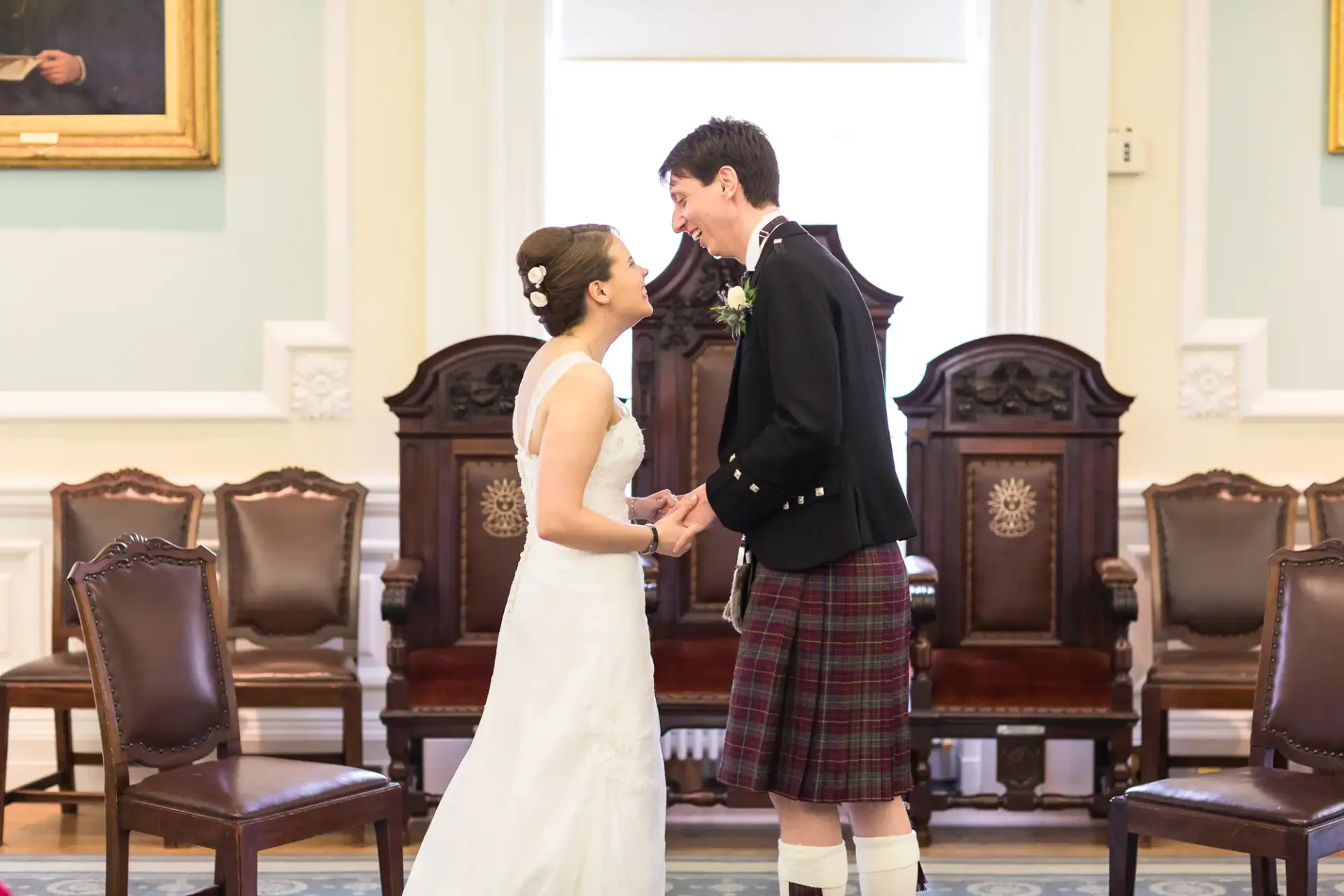A bride in a white dress and a groom in a kilt hold hands and smile at each other in a formal room with wood-paneled walls and ornate chairs.