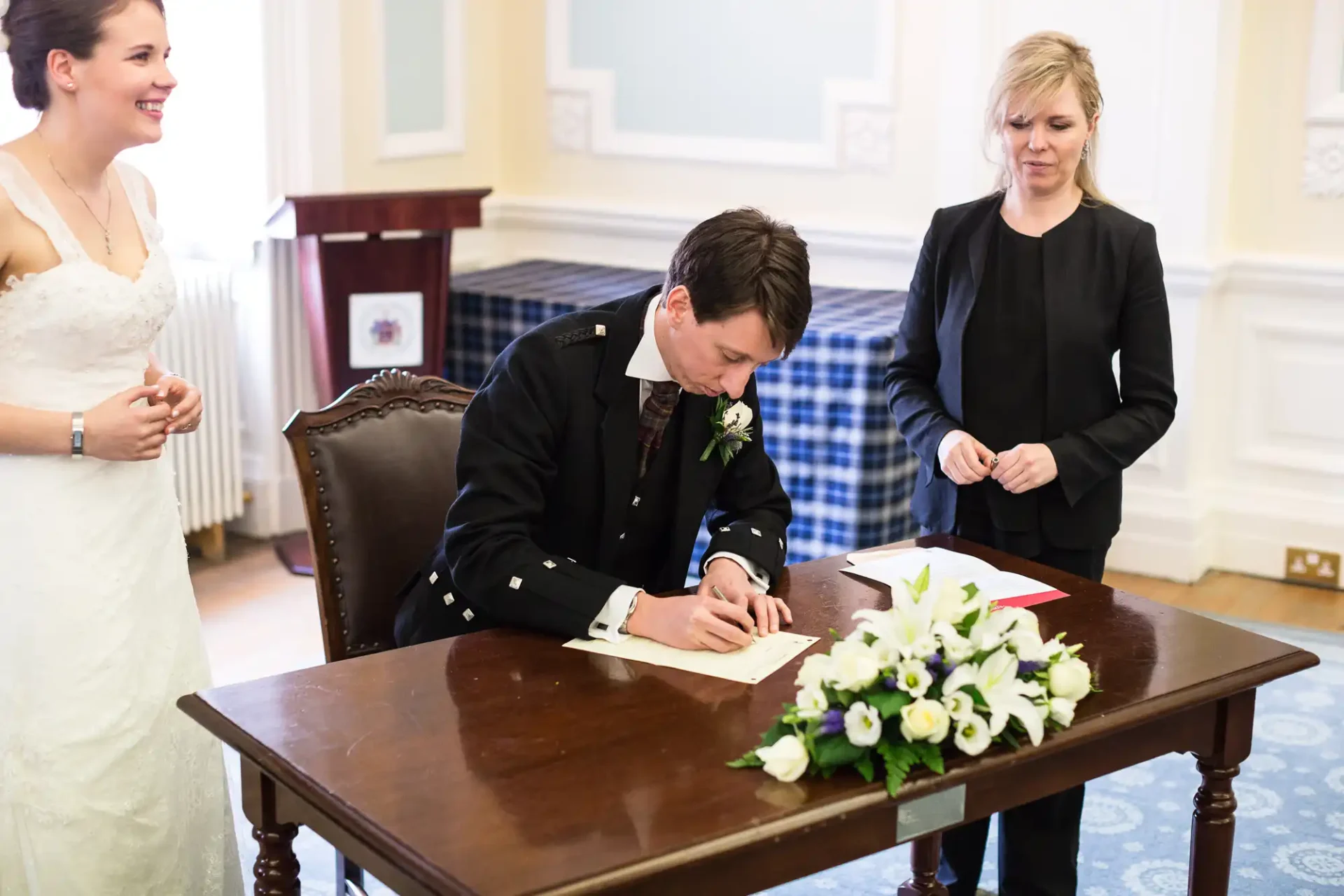 A groom in a dark suit signs a document at a table during a wedding ceremony, under the watchful eyes of a smiling bride and an officiant.