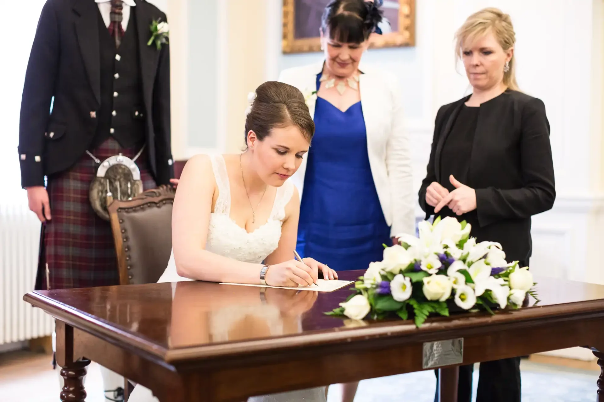 A bride in a white dress signs a document at a table, with a floral arrangement in front and two people watching her in a formal room.