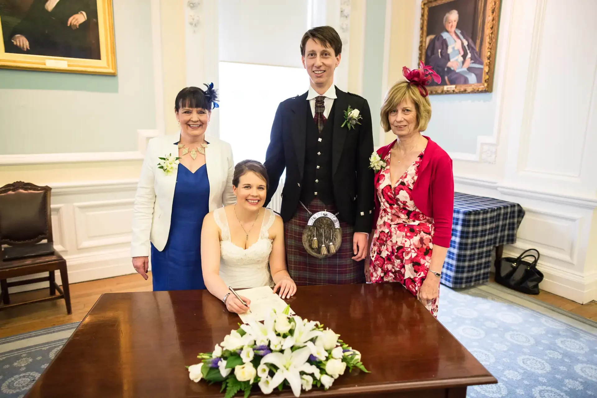 A bride seated at a table signing a document, flanked by a groom in a kilt and two smiling women in dresses, in an elegant room with portraits hanging.