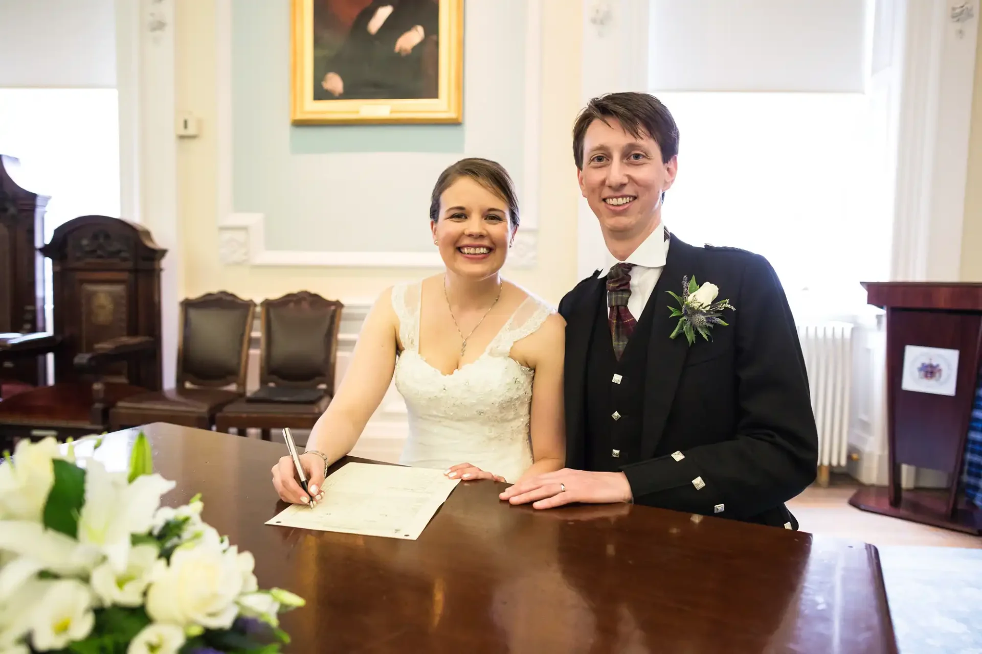 A smiling bride and groom signing a marriage certificate at a table in an elegant room with paintings and antique furniture.