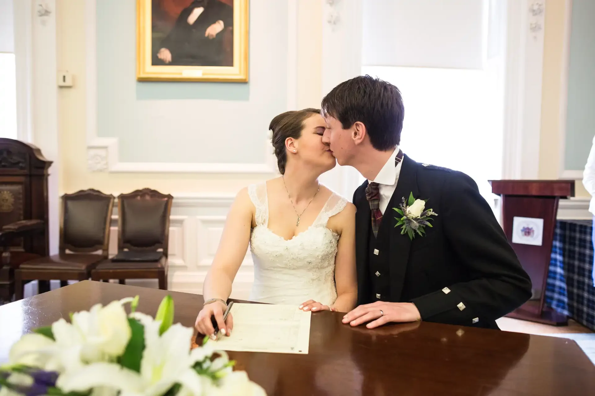 A newlywed couple kissing while signing their marriage certificate at a table, with a floral bouquet in the foreground and elegant room decor.