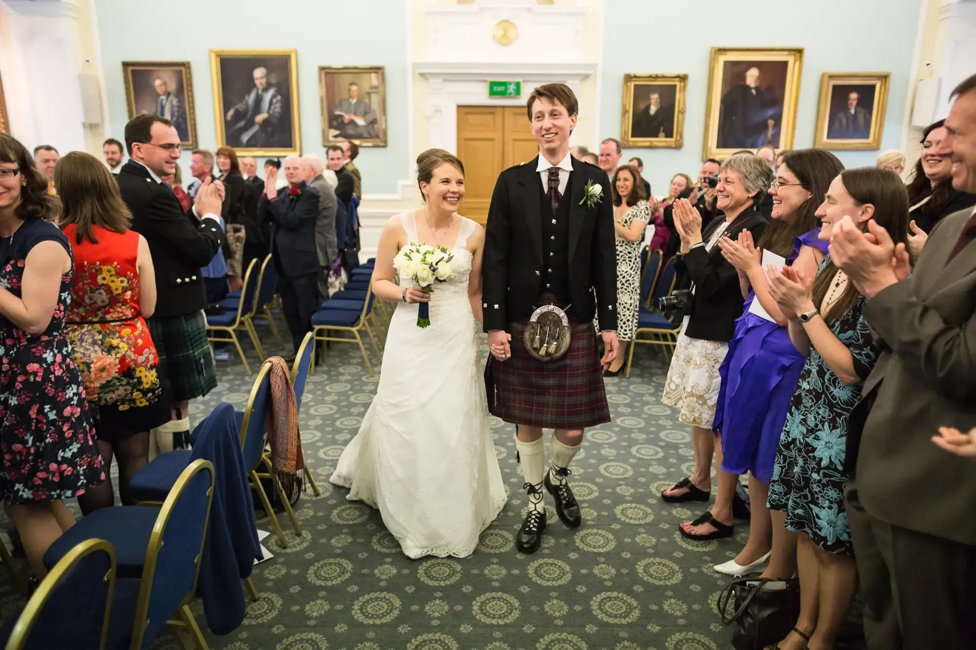 A newlywed couple smiling as they walk through a crowd of clapping guests in a hall adorned with portraits.