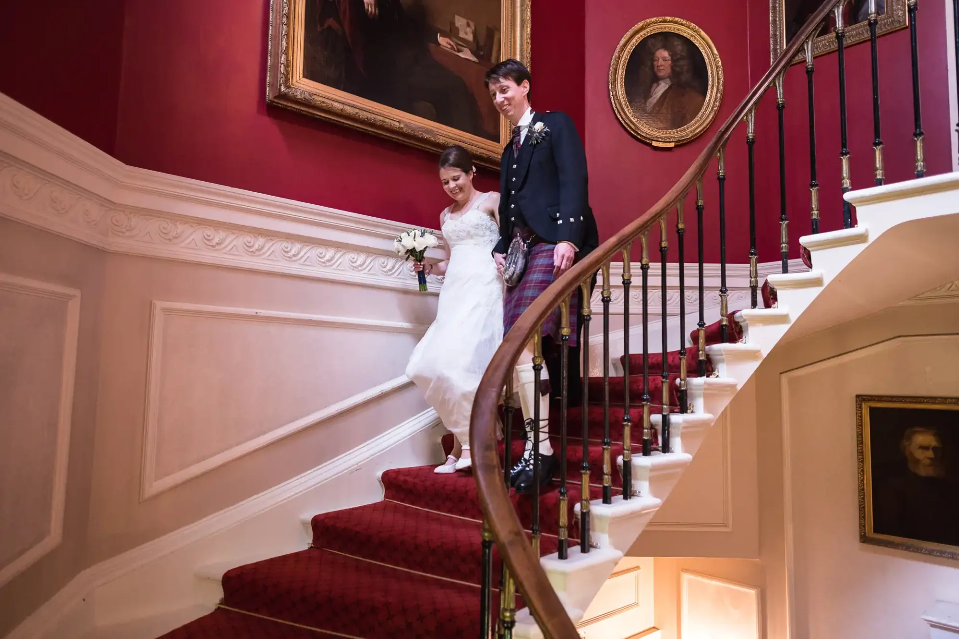 A bride and groom in traditional attire smiling as they descend a grand staircase with red carpeting in an elegant interior.