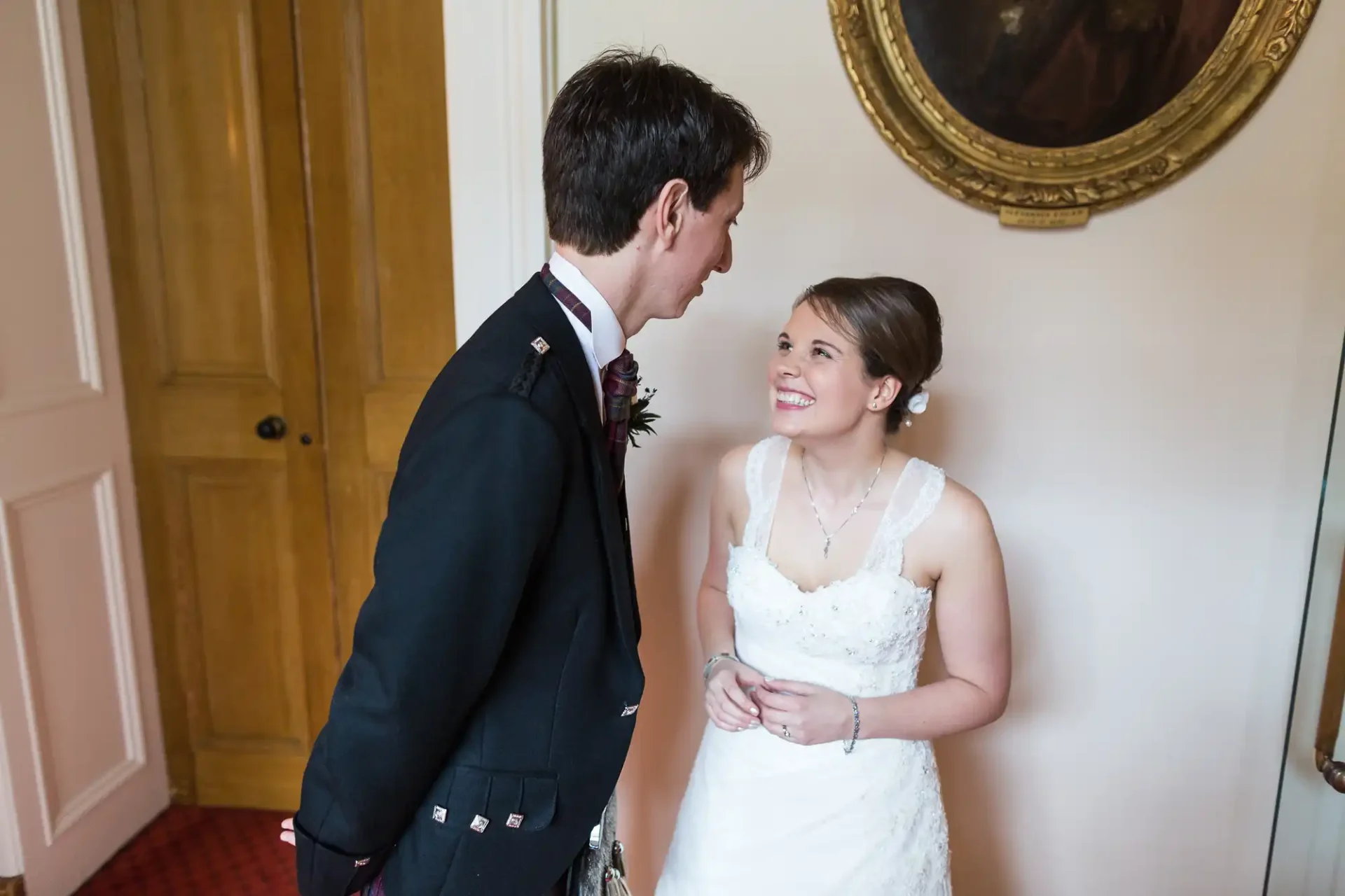 A bride in a white dress and a groom in a black suit share a joyful moment, smiling at each other indoors.