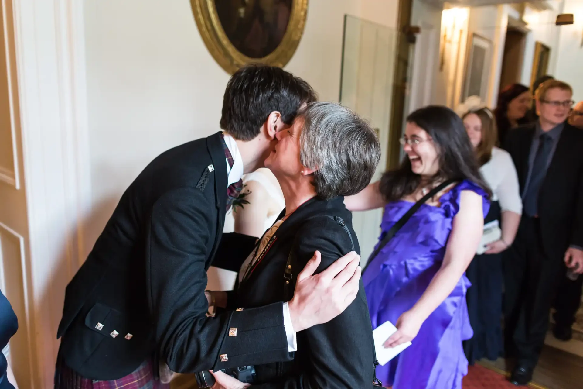 A couple in formal attire kissing each other on the cheek during a wedding ceremony, surrounded by smiling guests.