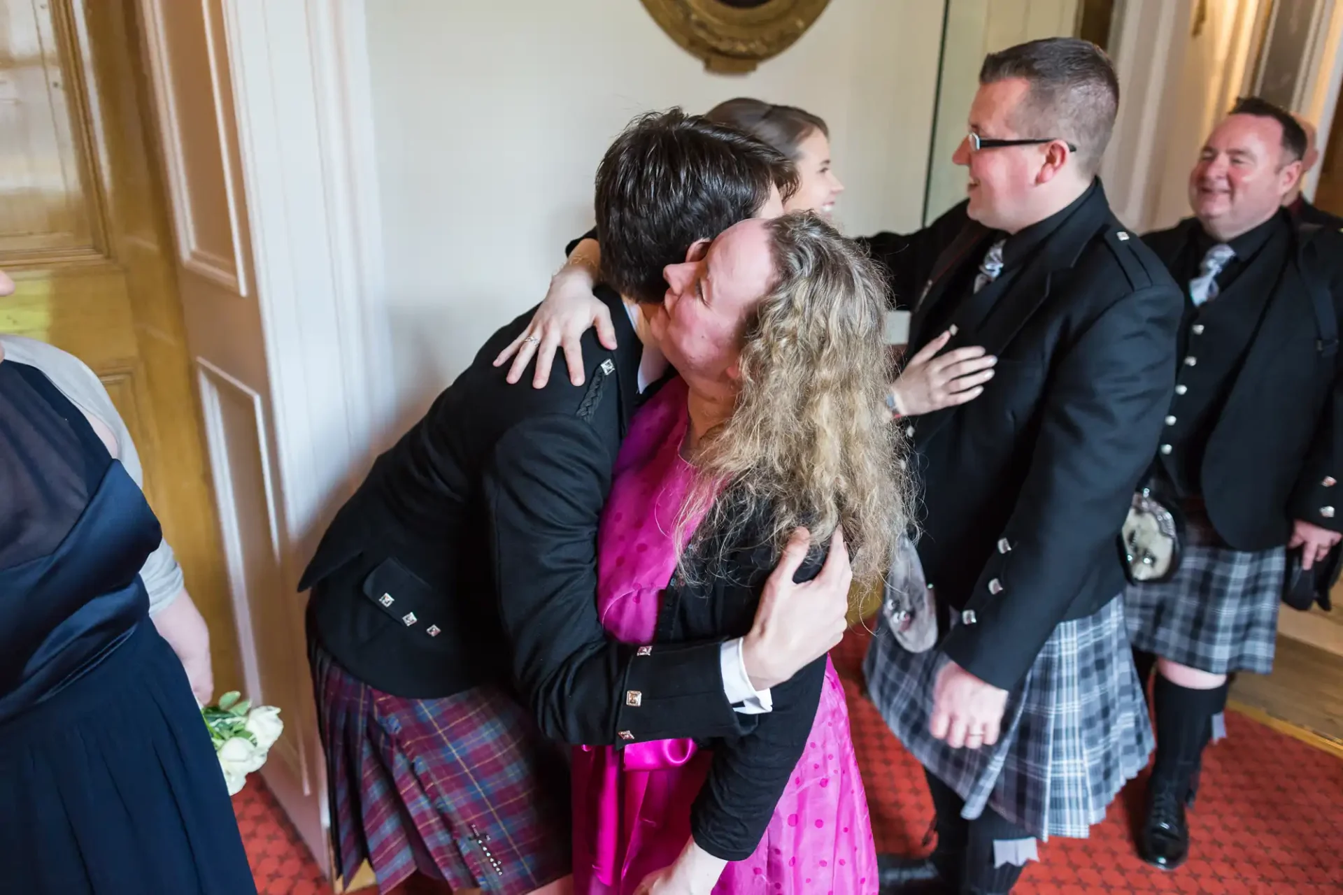 A man in a kilt hugs a woman in a pink dress, both smiling, in a room with others in formal attire, some also wearing kilts.