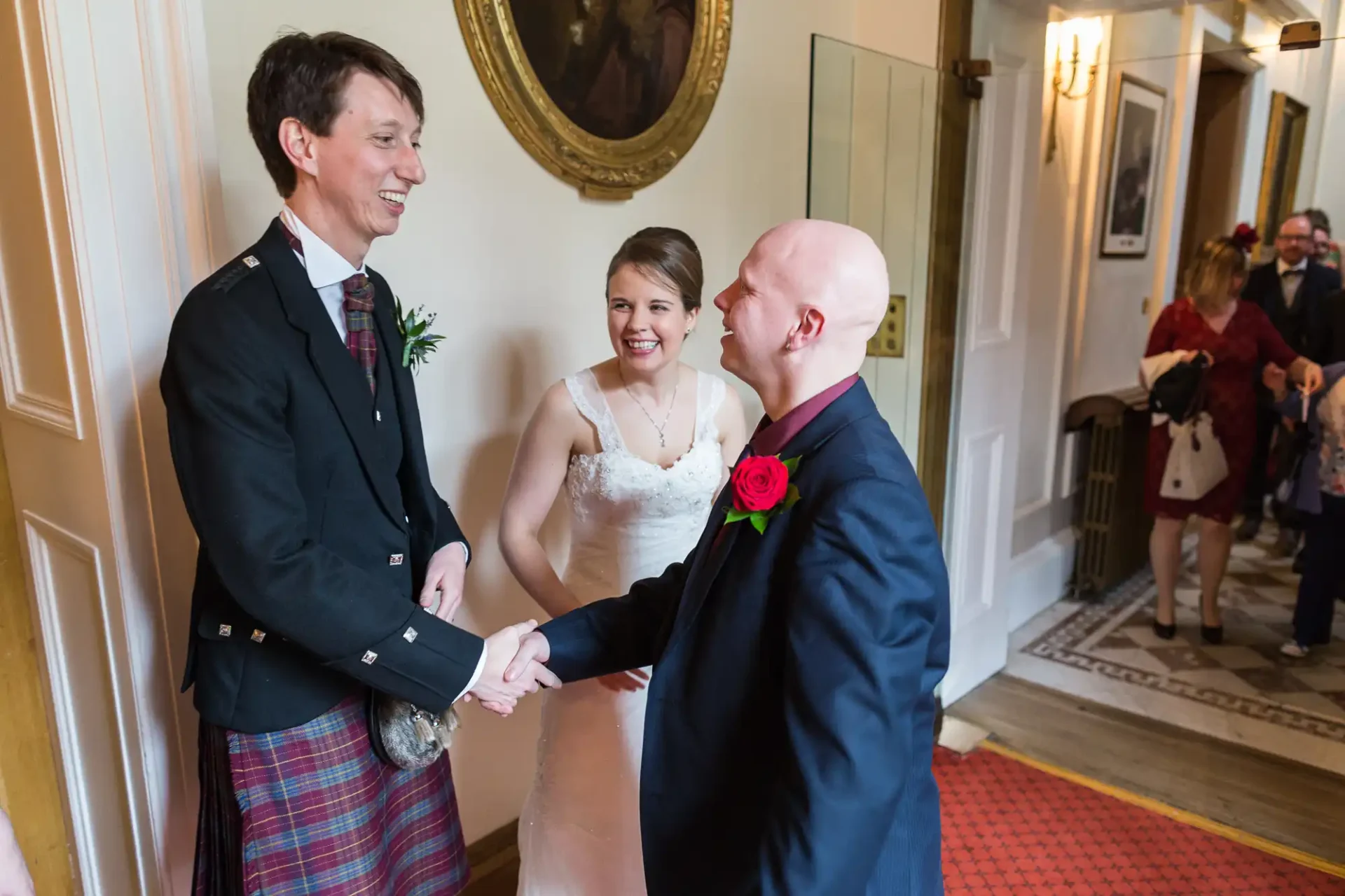 A bride and groom shake hands with a guest at their wedding, the groom in a kilt and the bride in a white dress, smiling in a hallway with onlookers.