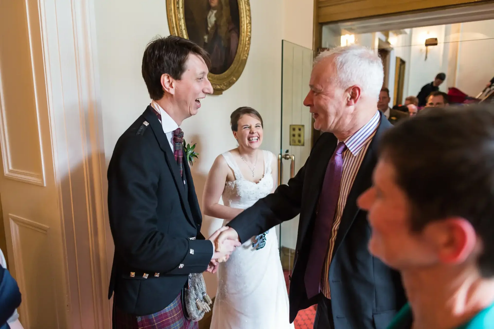 A joyful bride and groom shaking hands with an older man in a crowded room, with guests and laughter in the background.