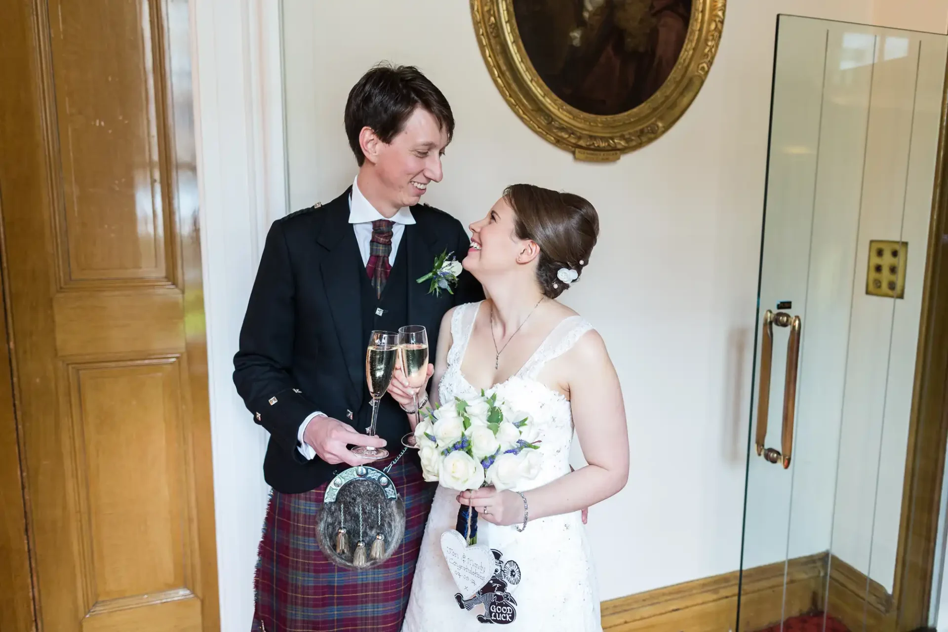 A couple in wedding attire smiling at each other, holding champagne flutes, with the groom wearing a kilt.