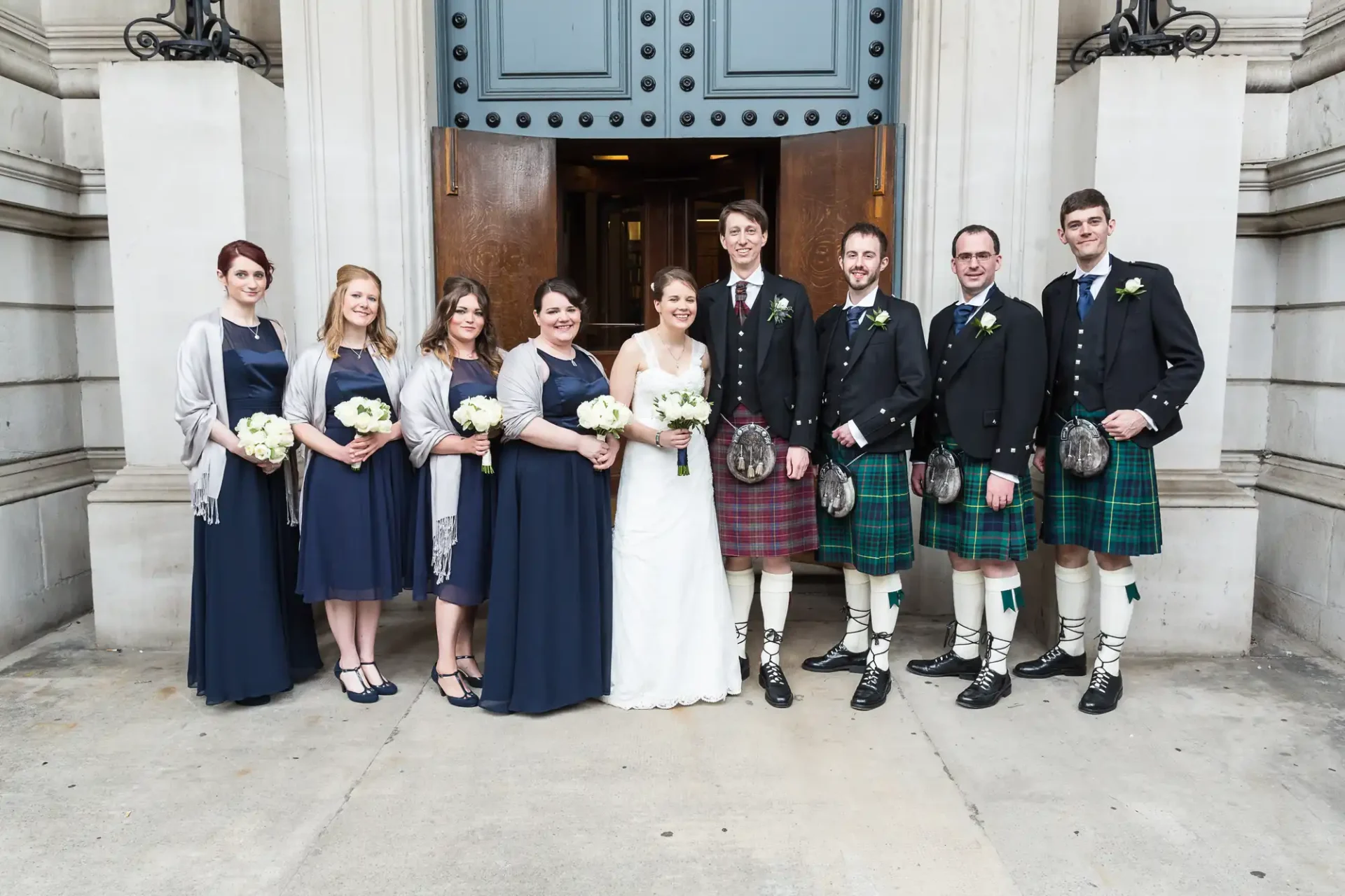 A wedding group posing in front of a building, with the bride and groom flanked by bridesmaids in blue and groomsmen in kilts.