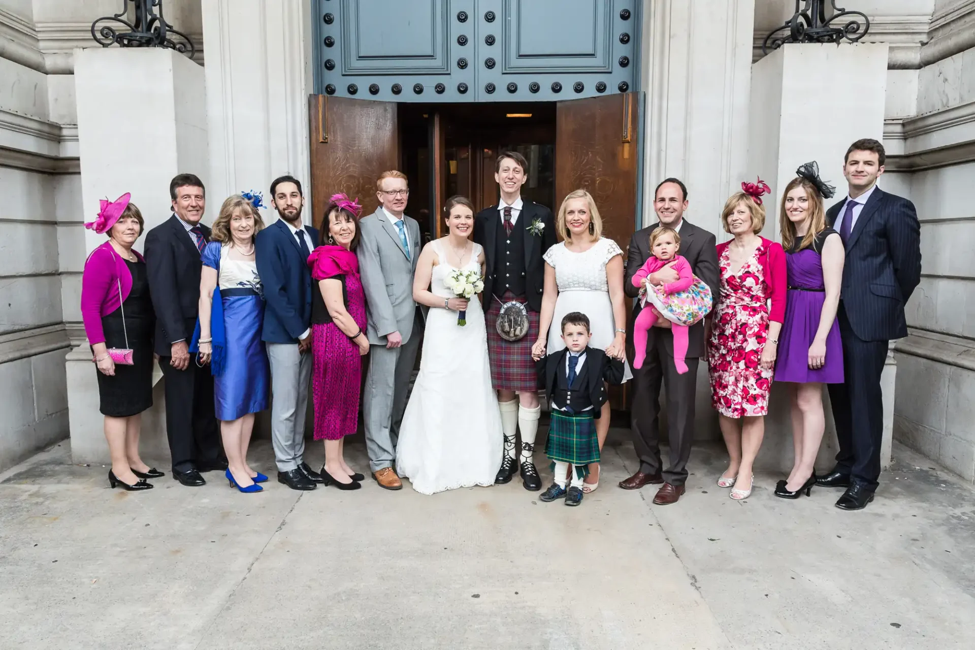 A group of family members in formal attire posing for a photo at a wedding outside a building entrance. some wear kilts; others have hats.