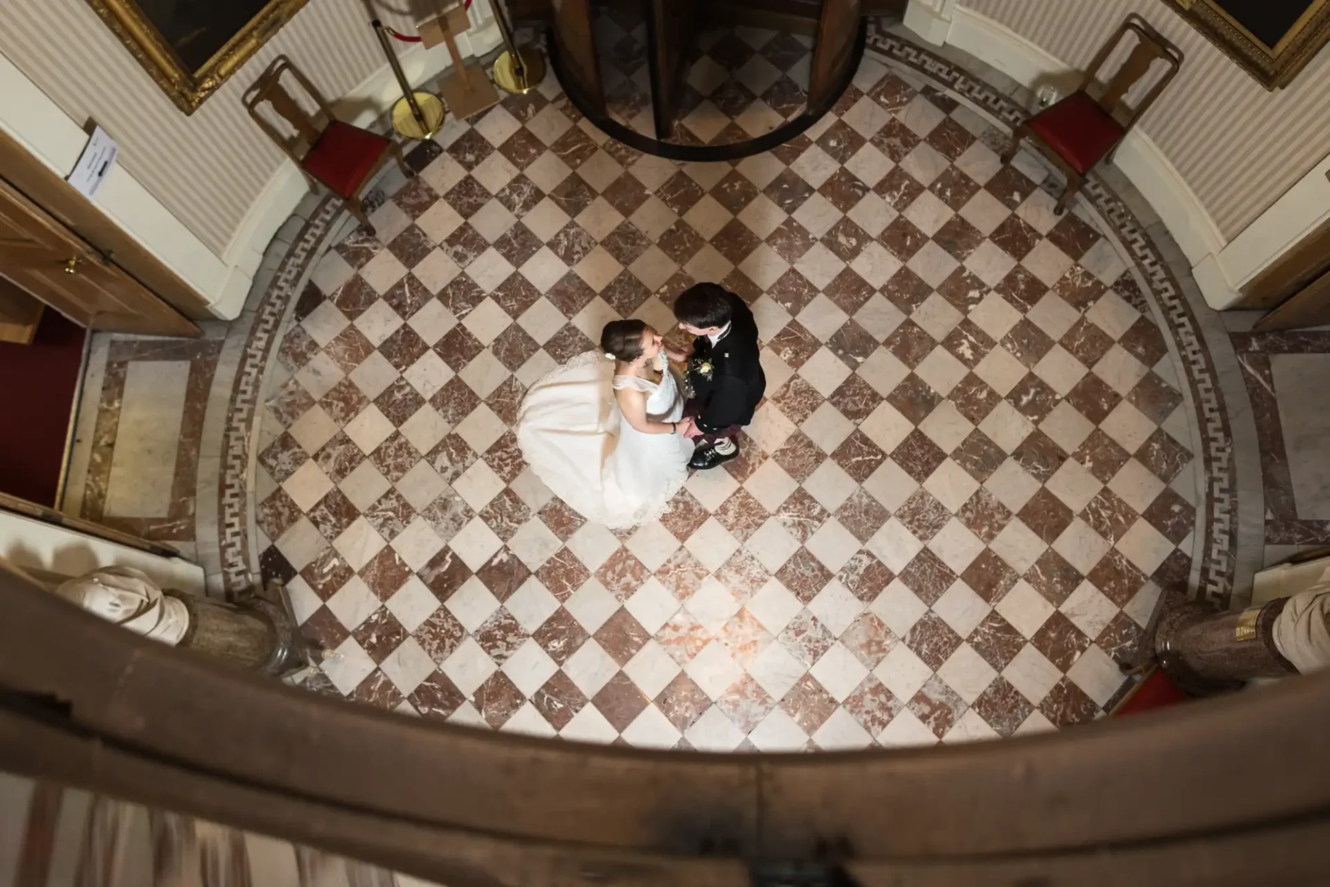 Aerial view of a bride and groom sitting on the floor, holding hands in a circular room with checkered flooring.