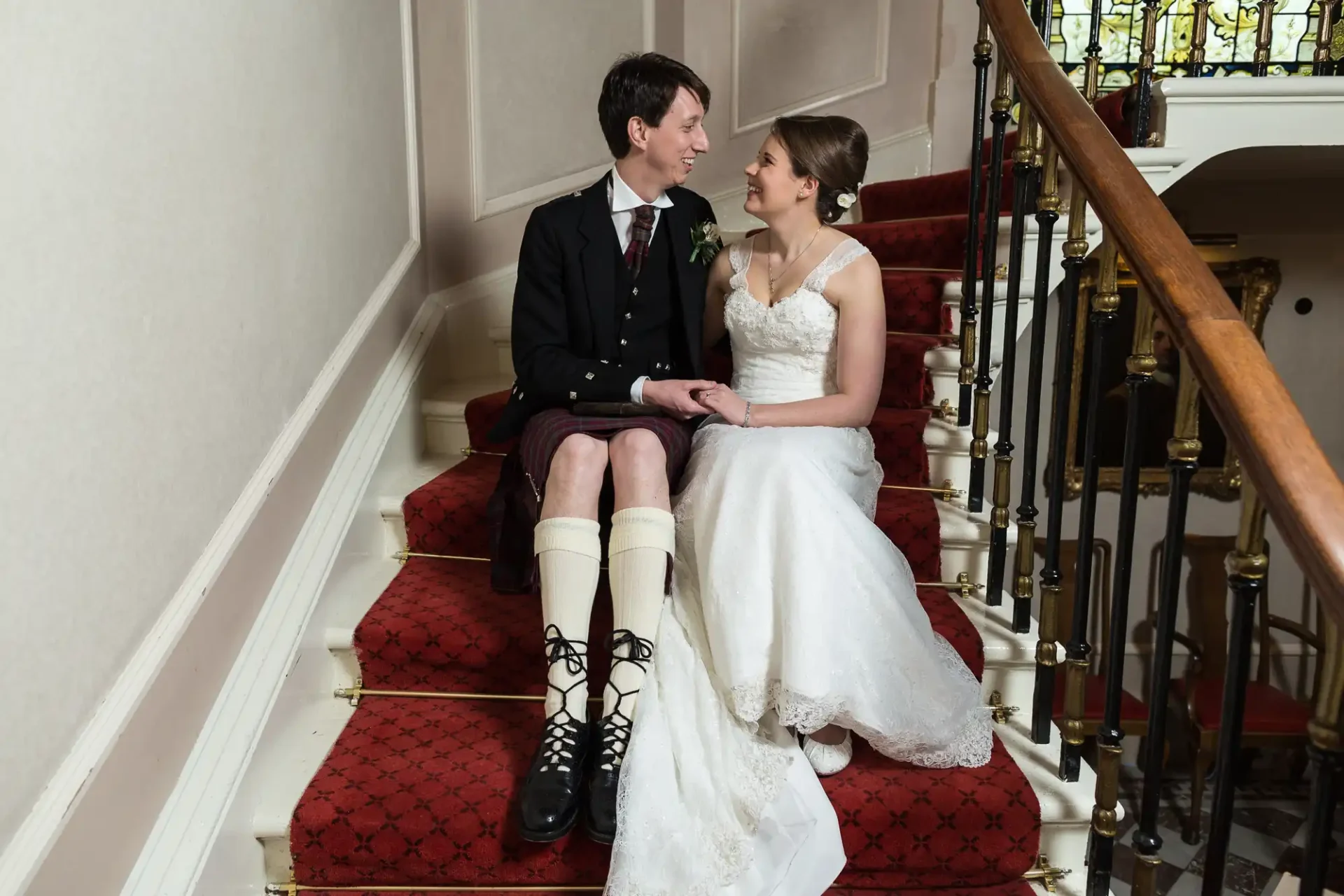 Newlyweds sitting on a red carpeted staircase, exchanging smiles. the groom is wearing a kilt and sporran, while the bride is in a white strapless dress.