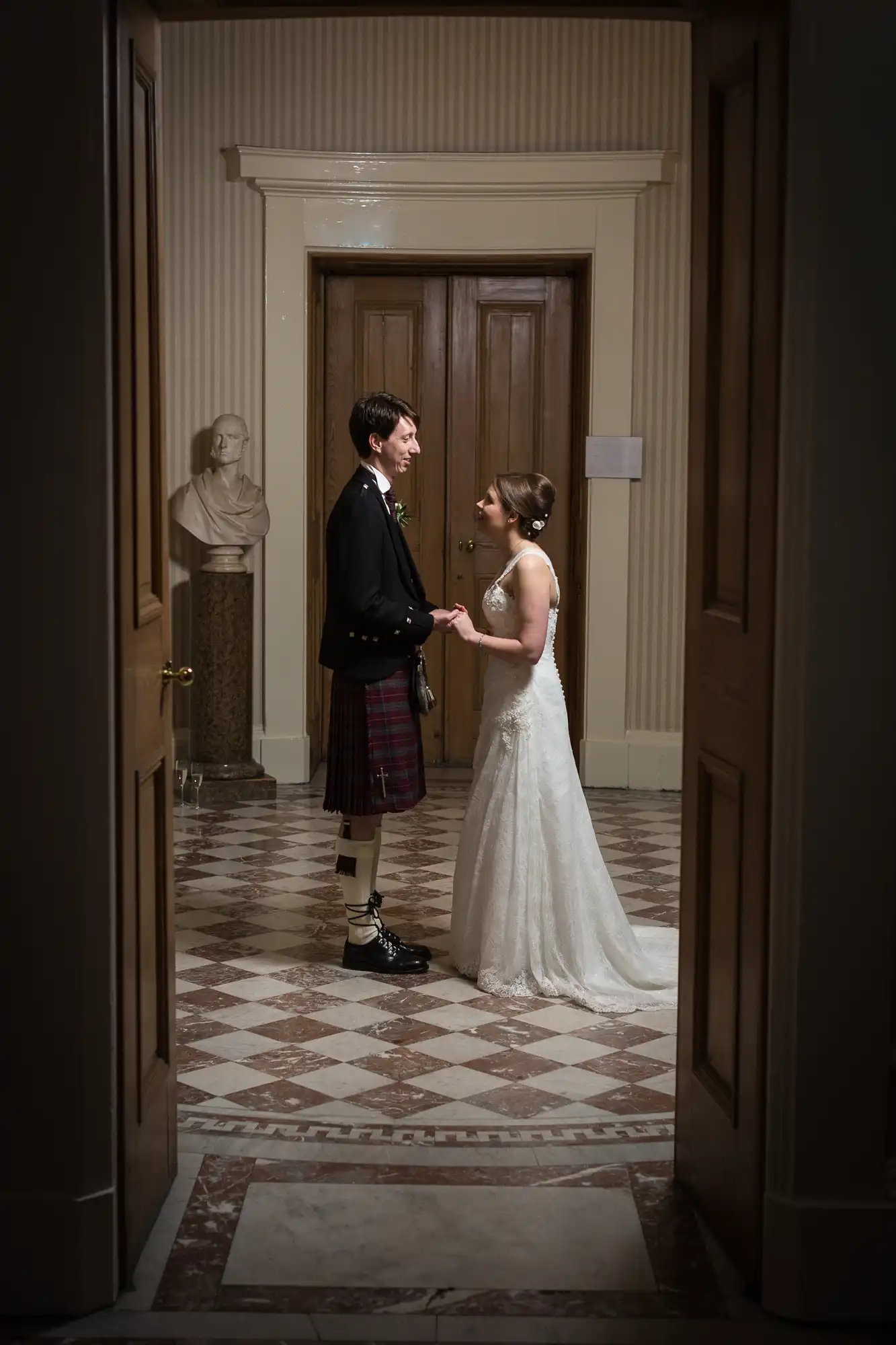 A bride and groom in formal attire smiling at each other in a hallway, the groom wearing a kilt and the bride in a lace gown.