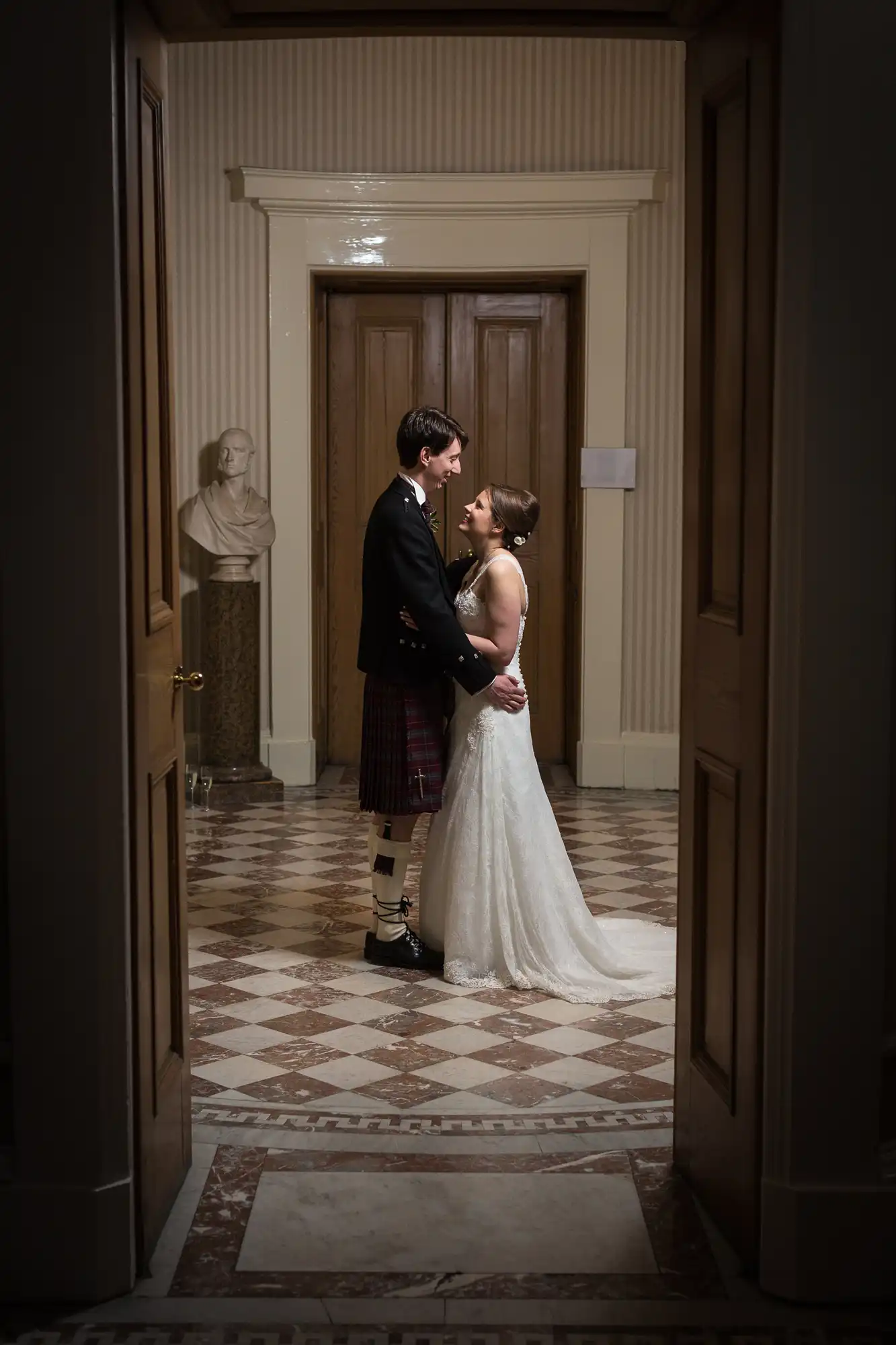 A bride in a white gown and a groom in a kilt share an intimate moment in a dimly lit hallway with classical decor.