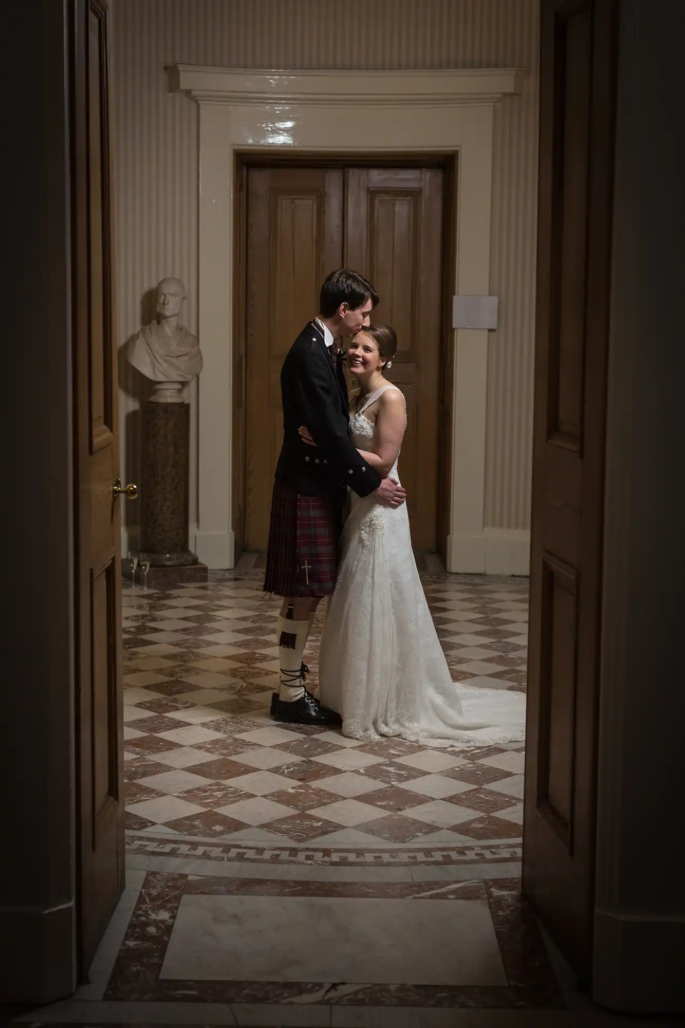 A bride and groom embracing in a hallway, the groom wearing a kilt and the bride in a lace gown, surrounded by elegant architectural details.