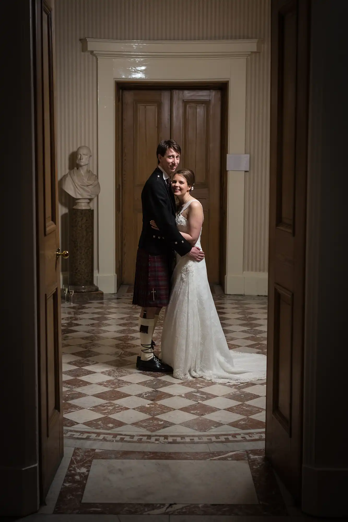 A bride and groom embracing in a hallway, the groom in a kilt and the bride in a lace dress, with warm lighting highlighting them from the front.