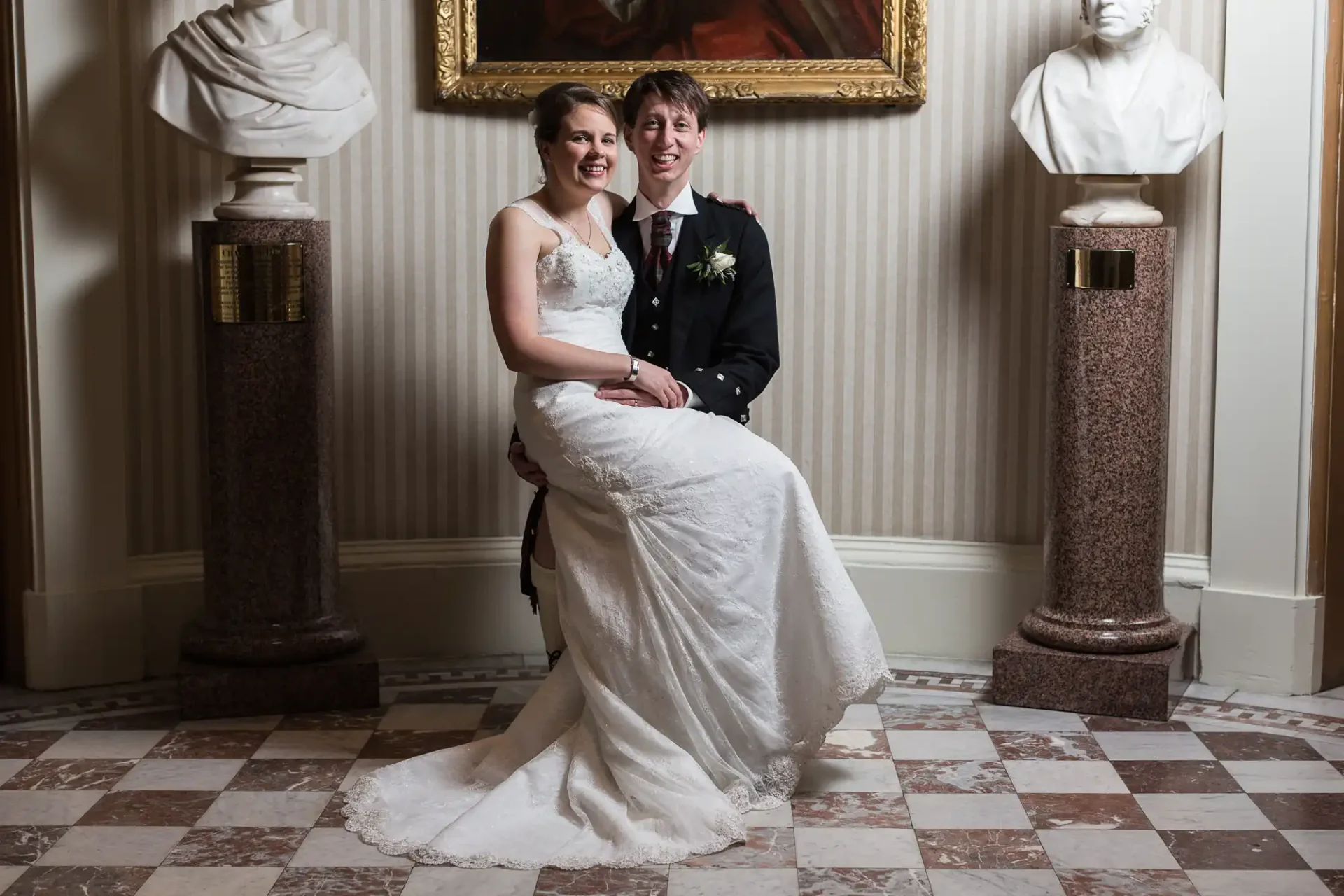 A bride and groom smiling, seated together between two classical bust sculptures in an elegant room with a checkered floor and framed artwork.