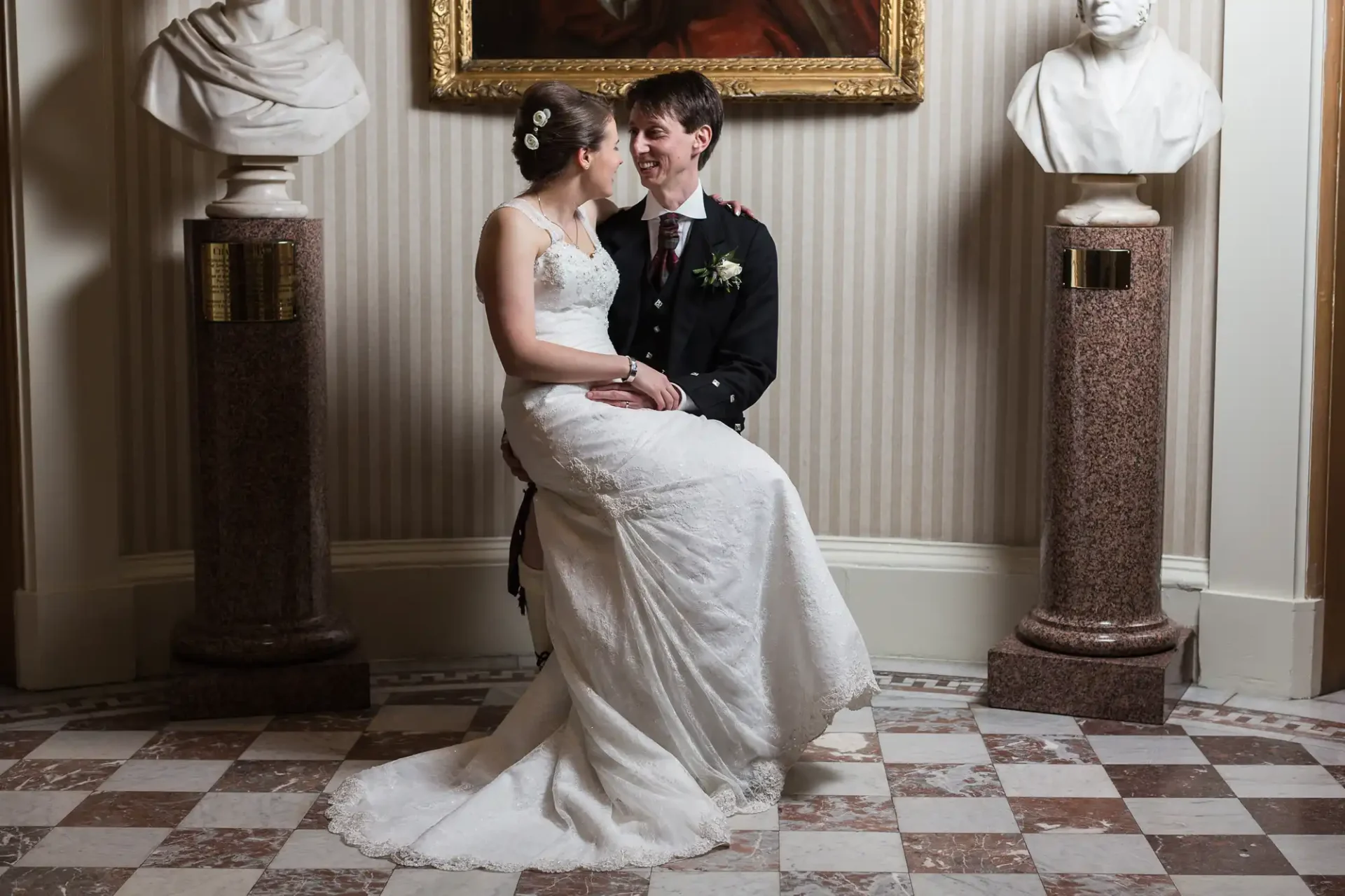 A bride in a white gown and a groom in a black suit seated closely together, smiling, in a classic room with marble columns and bust sculptures.