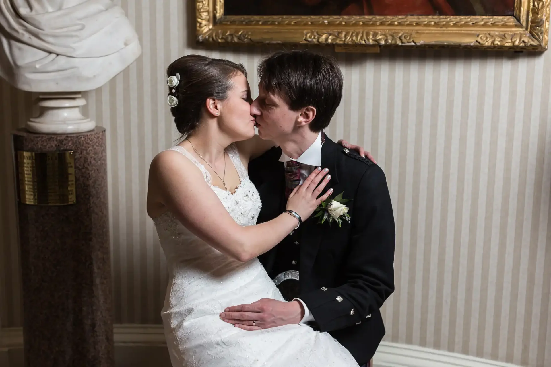 A bride and groom sharing a kiss, with the bride seated on the groom's lap, both dressed in formal wedding attire, in a room with elegant decor.