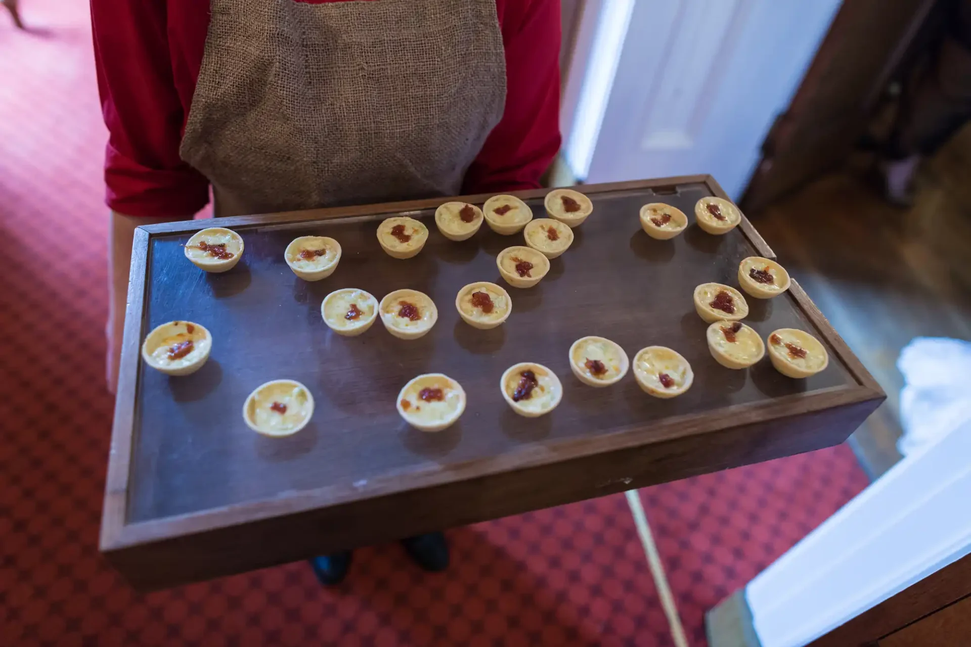 A person holding a wooden tray filled with small, open-faced tarts in a room with a red carpet.